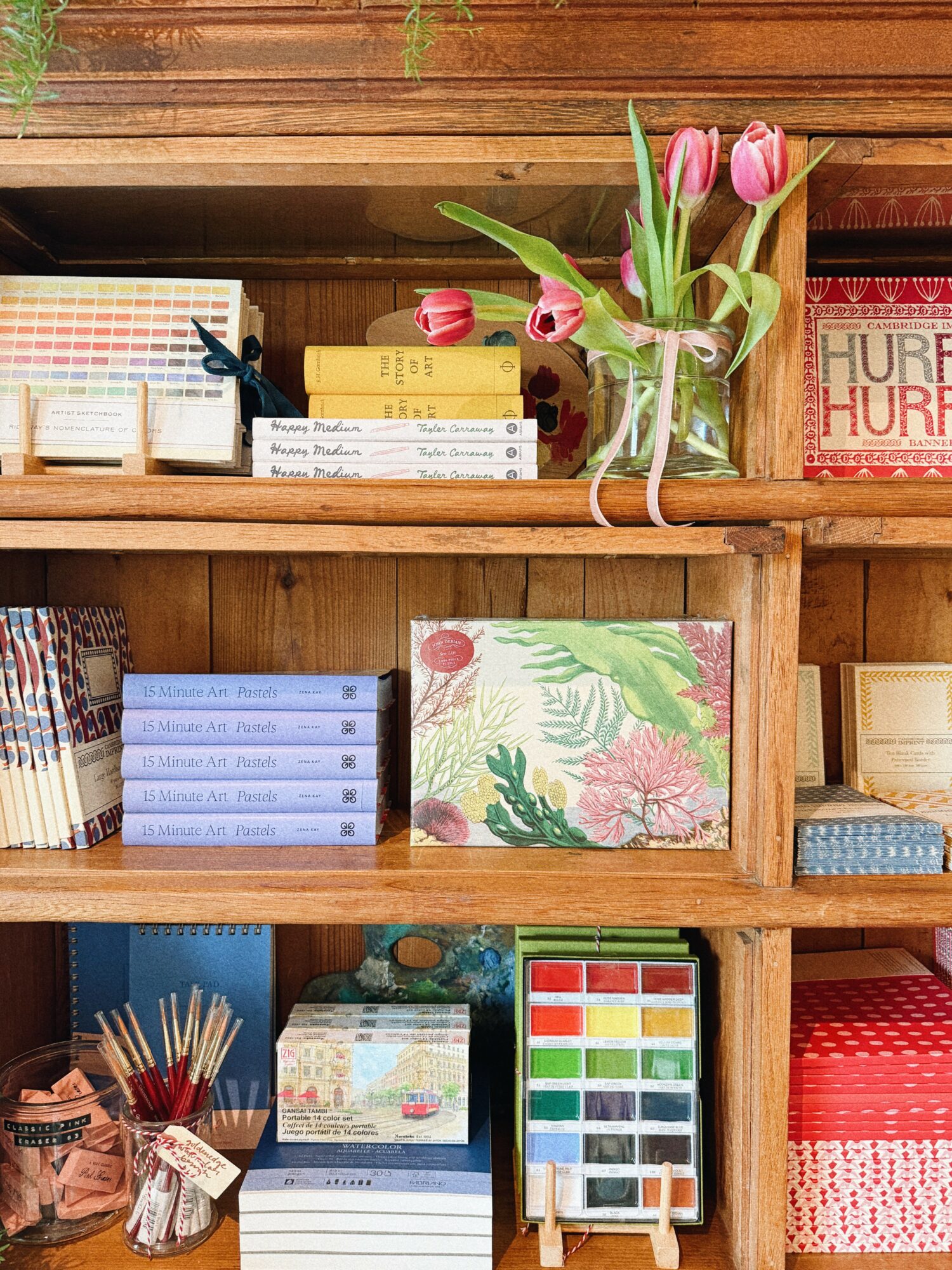 Wooden shelf with books, a vase of pink tulips, and decorative items, including a framed floral artwork and colorful boxes.