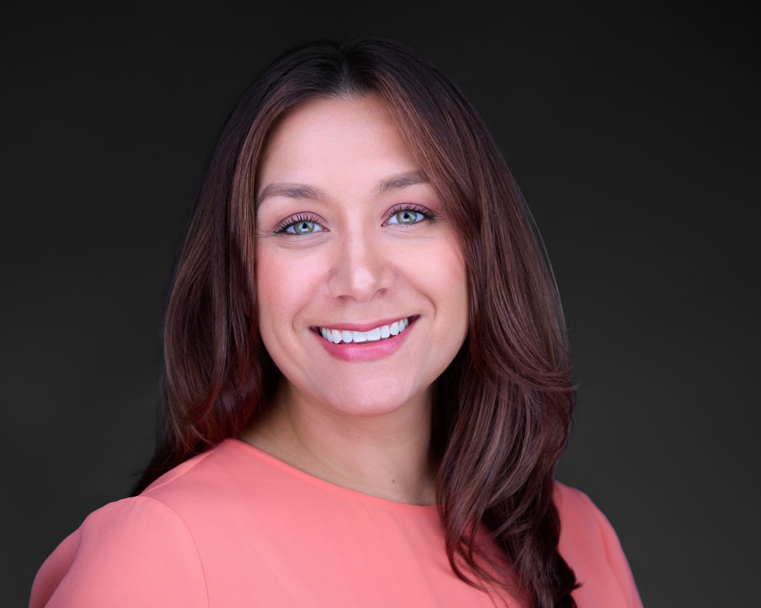 Portrait of a smiling woman with shoulder-length brown hair, wearing a pink top, against a dark background.