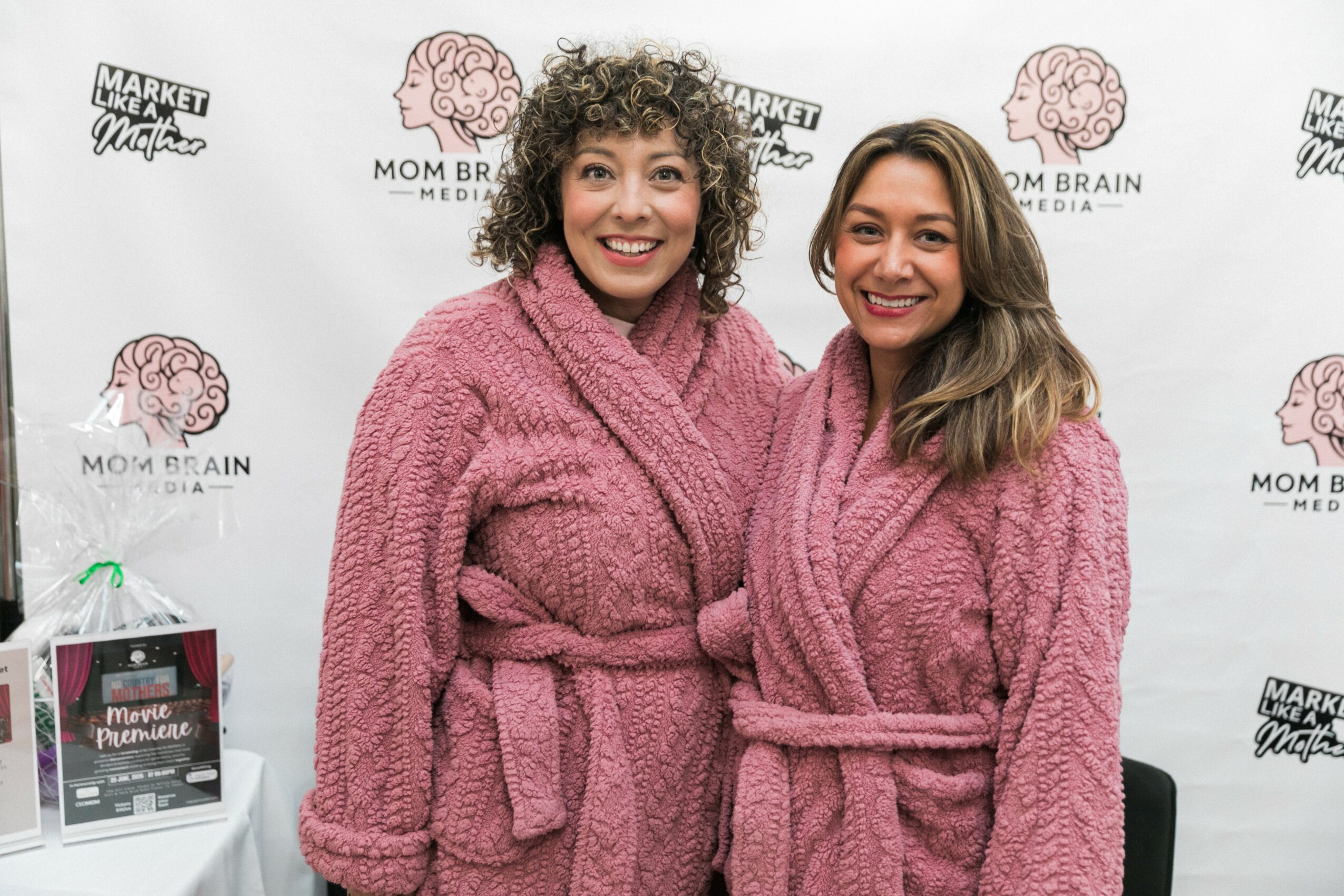 Two women in pink robes smiling in front of a backdrop with logos, one with curly hair, the other with straight hair.