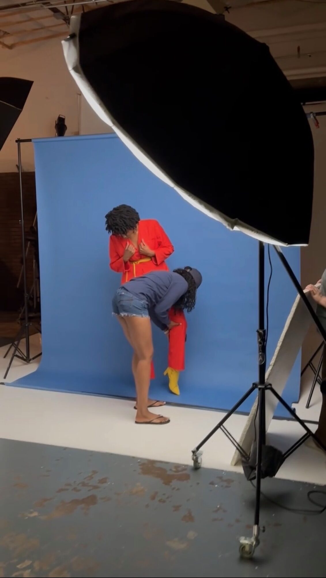 Person in red shirt and blue shorts bending over, with a blue backdrop and studio lighting equipment visible.
