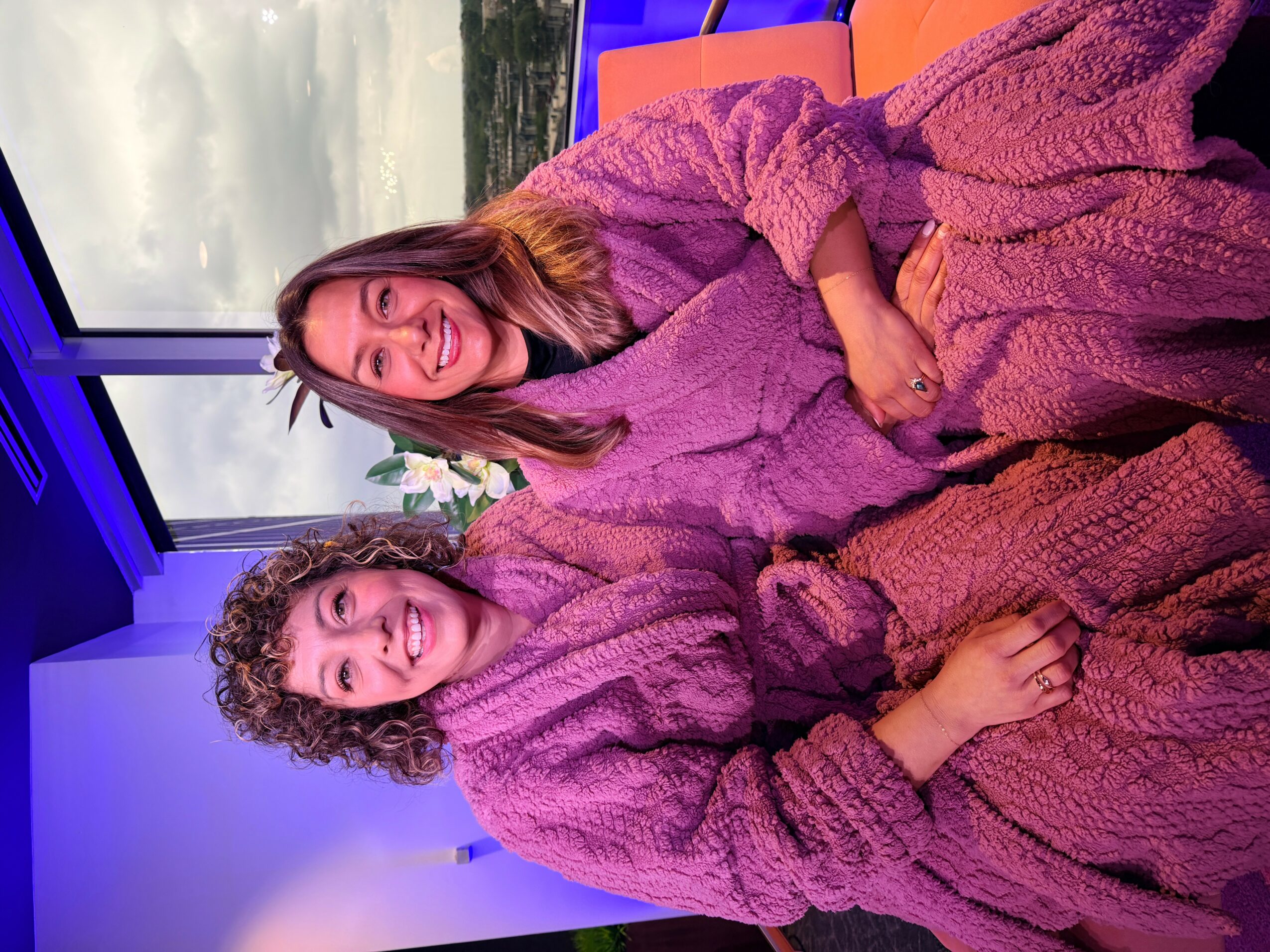 Two women sitting together, smiling, wearing matching purple robes, indoors near a window with a view outside.