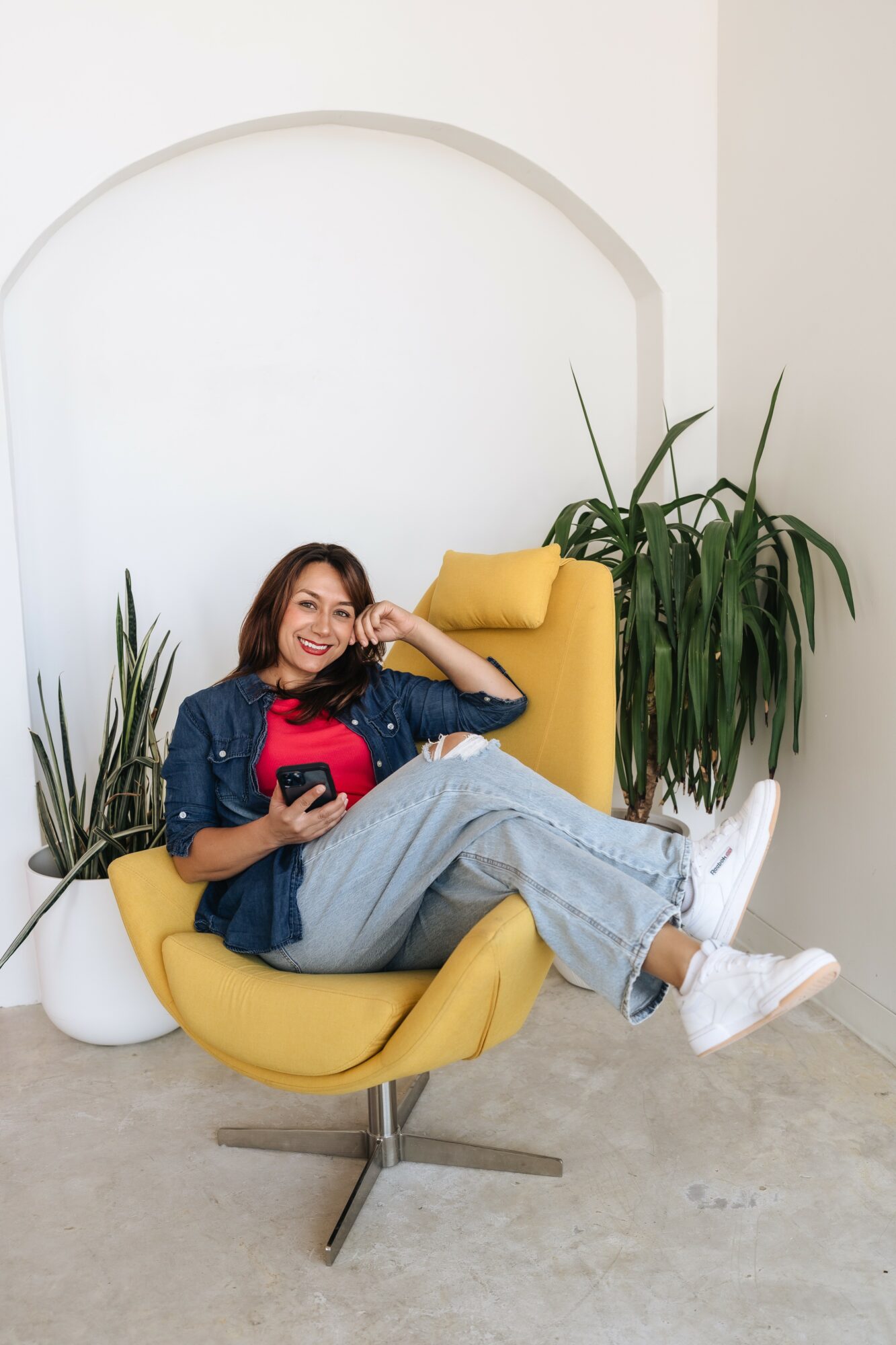Woman sitting on a yellow chair, holding a phone, smiling, with large green plants nearby, in a bright room.