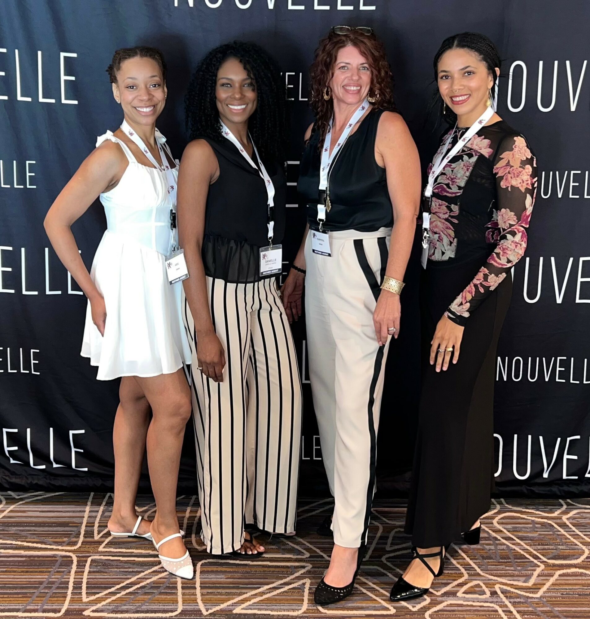 Four women standing together in front of a black backdrop with white text, smiling, dressed in stylish outfits.