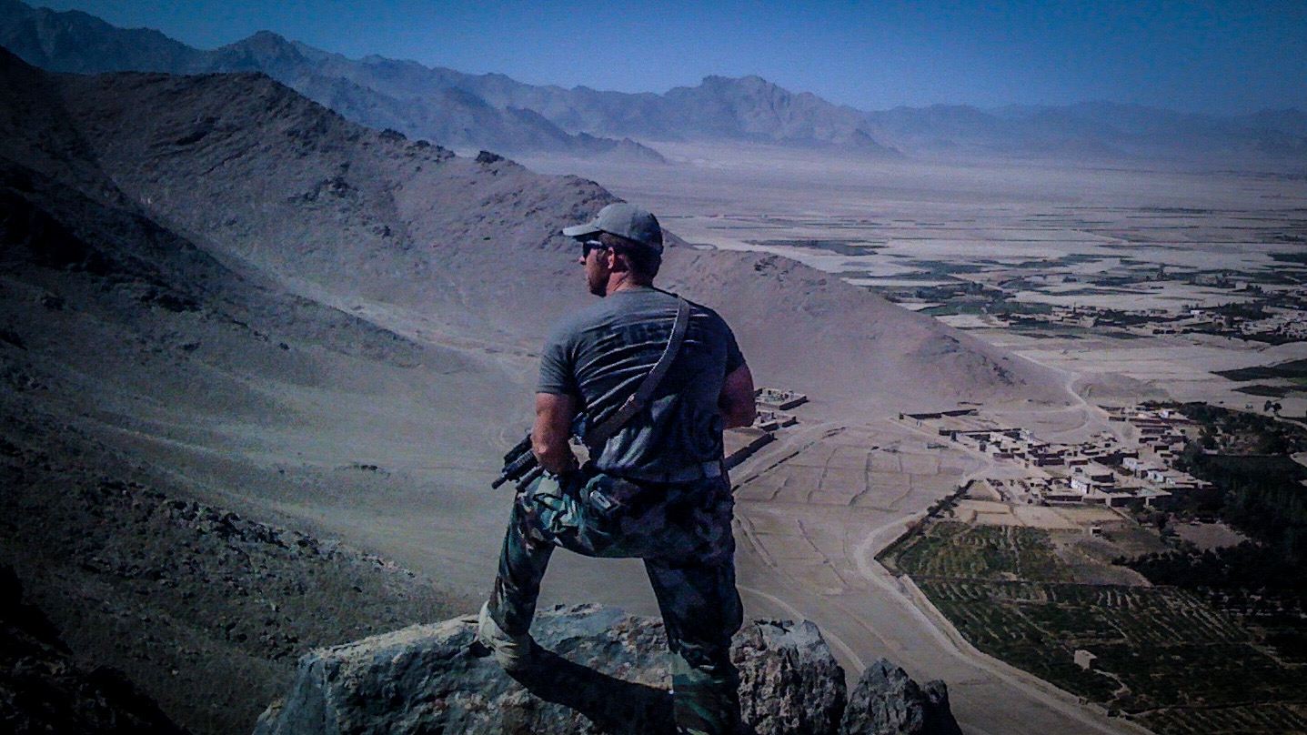 Person in camouflage clothing sitting on a rock overlooking a desert landscape with mountains in the distance.