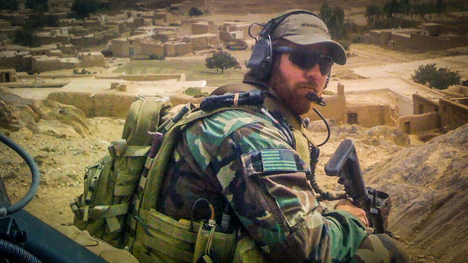 Soldier in camouflage uniform with backpack and headset, holding a rifle, in a desert-like environment with buildings in the background.