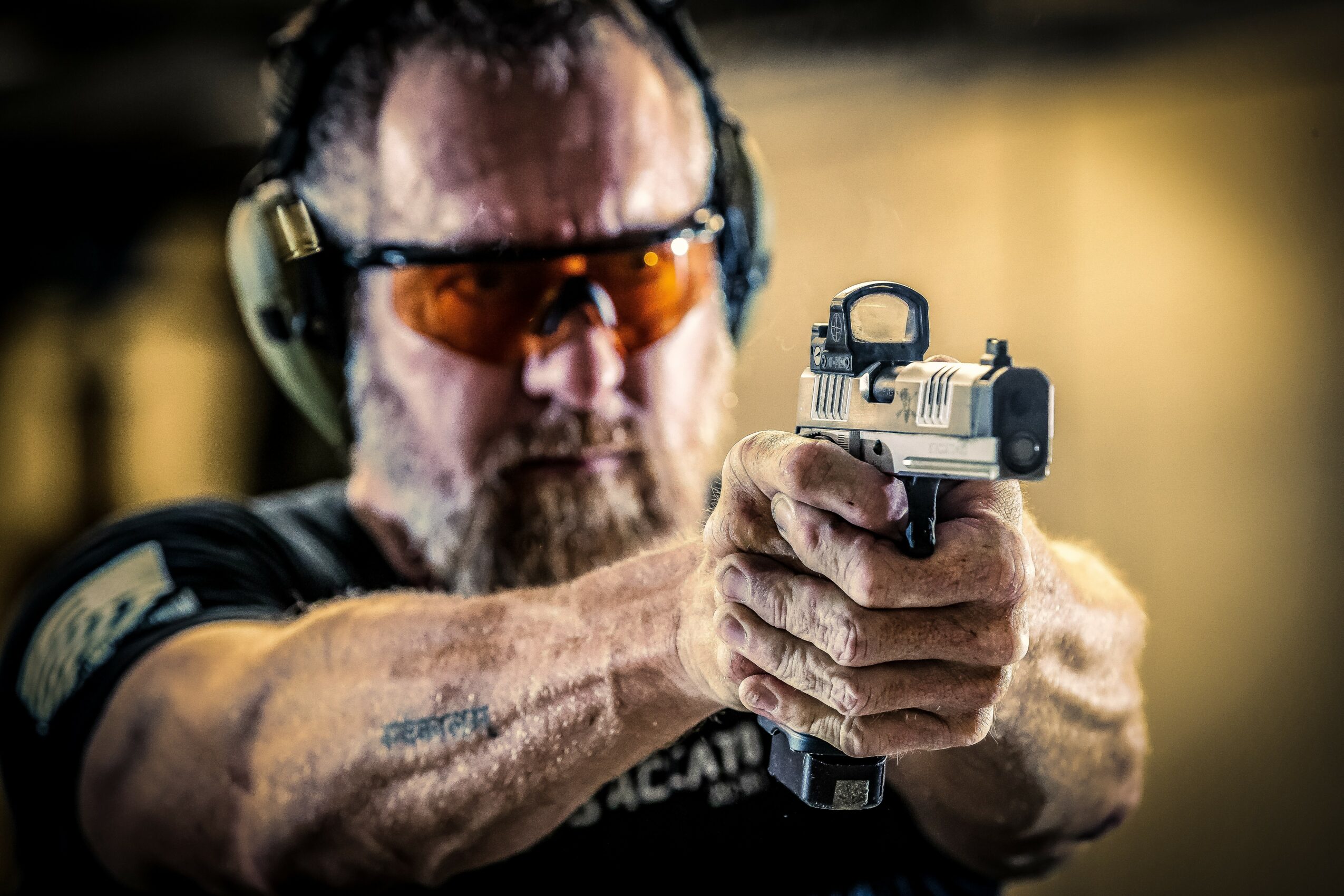 Man aiming a handgun, wearing protective glasses and ear protection, in a shooting range, with a blurred background.