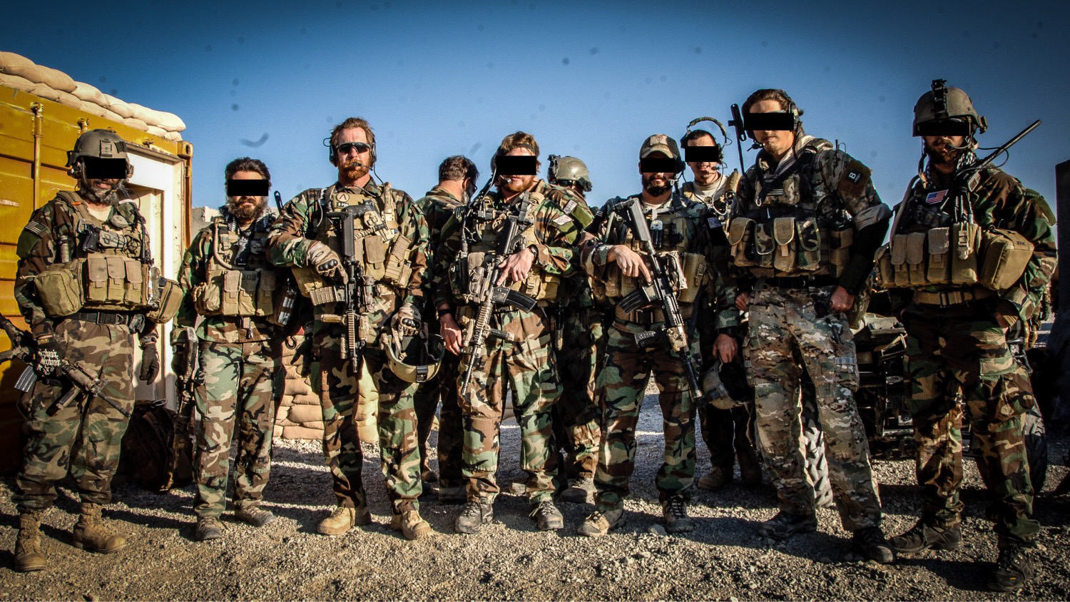 Group of soldiers in camouflage uniforms standing outdoors, some wearing helmets and tactical gear, against a blue sky.