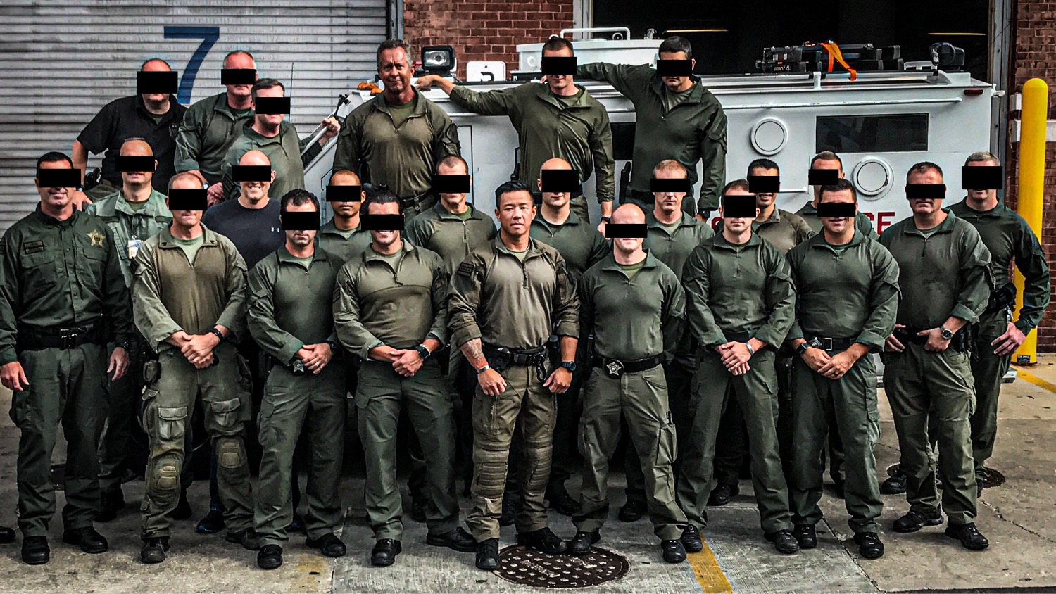 Group of uniformed personnel standing outdoors in front of a large vehicle, some wearing masks, posing for a photo.