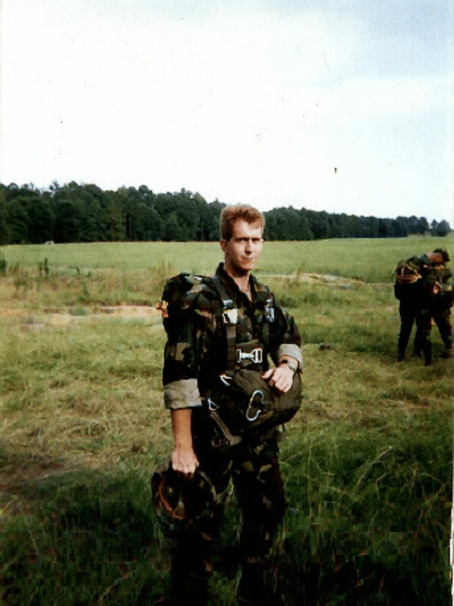 Soldier in camouflage uniform standing in grassy field with other soldiers in background, trees, and sky.