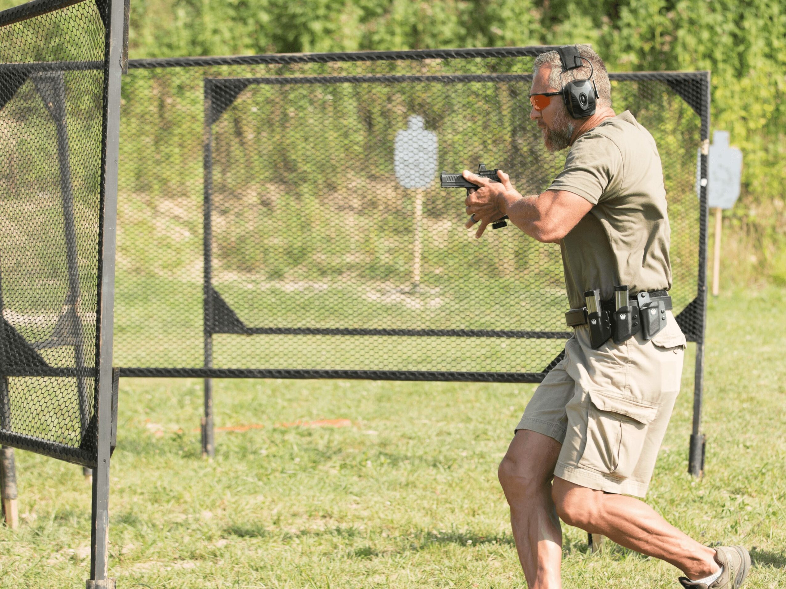 Man wearing headphones and sunglasses aiming a handgun at a shooting range outdoors, with targets in the background.