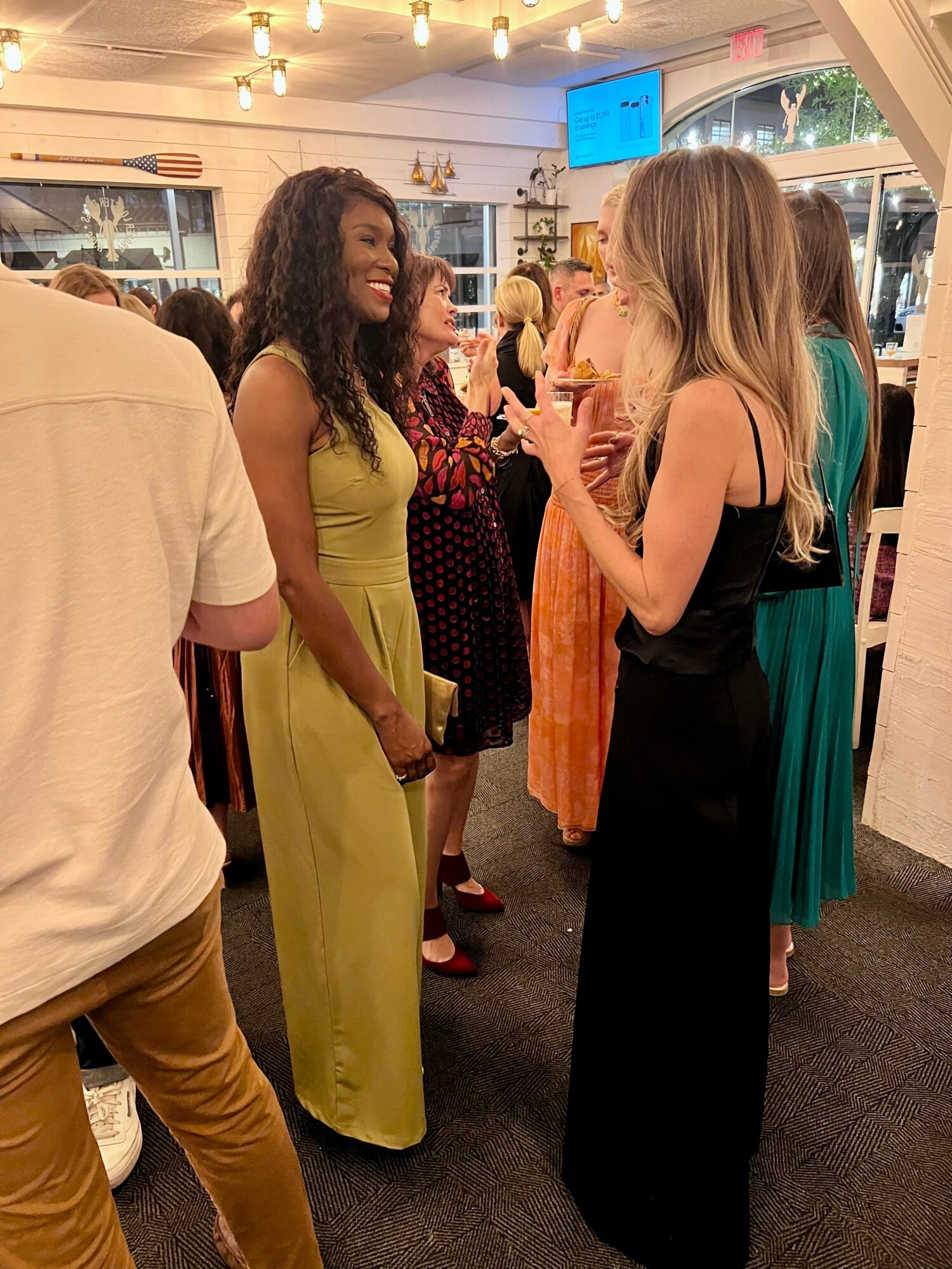 Two women are talking and smiling in a lively indoor setting with other people in the background.