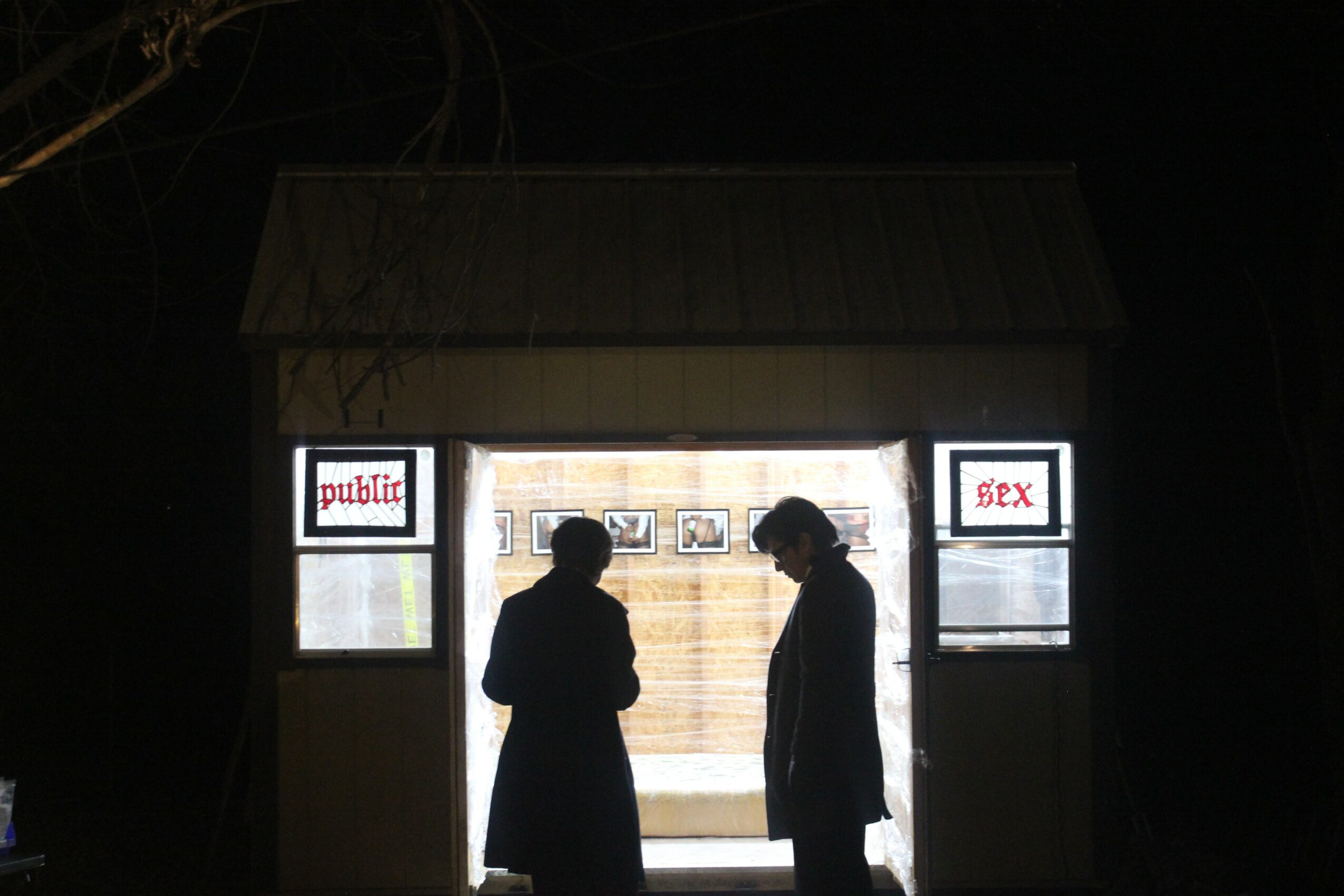 Silhouettes of two people standing in front of a brightly lit structure with windows, at night.