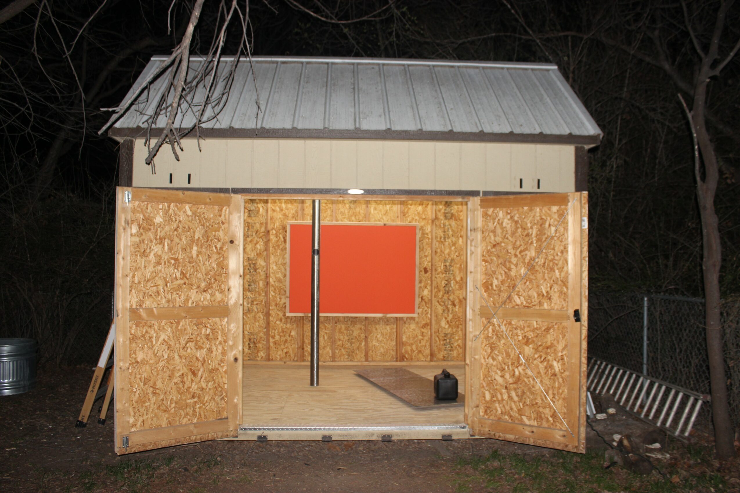 Small wooden structure with open doors, illuminated interior, and a red panel on the back wall, outdoors at night.