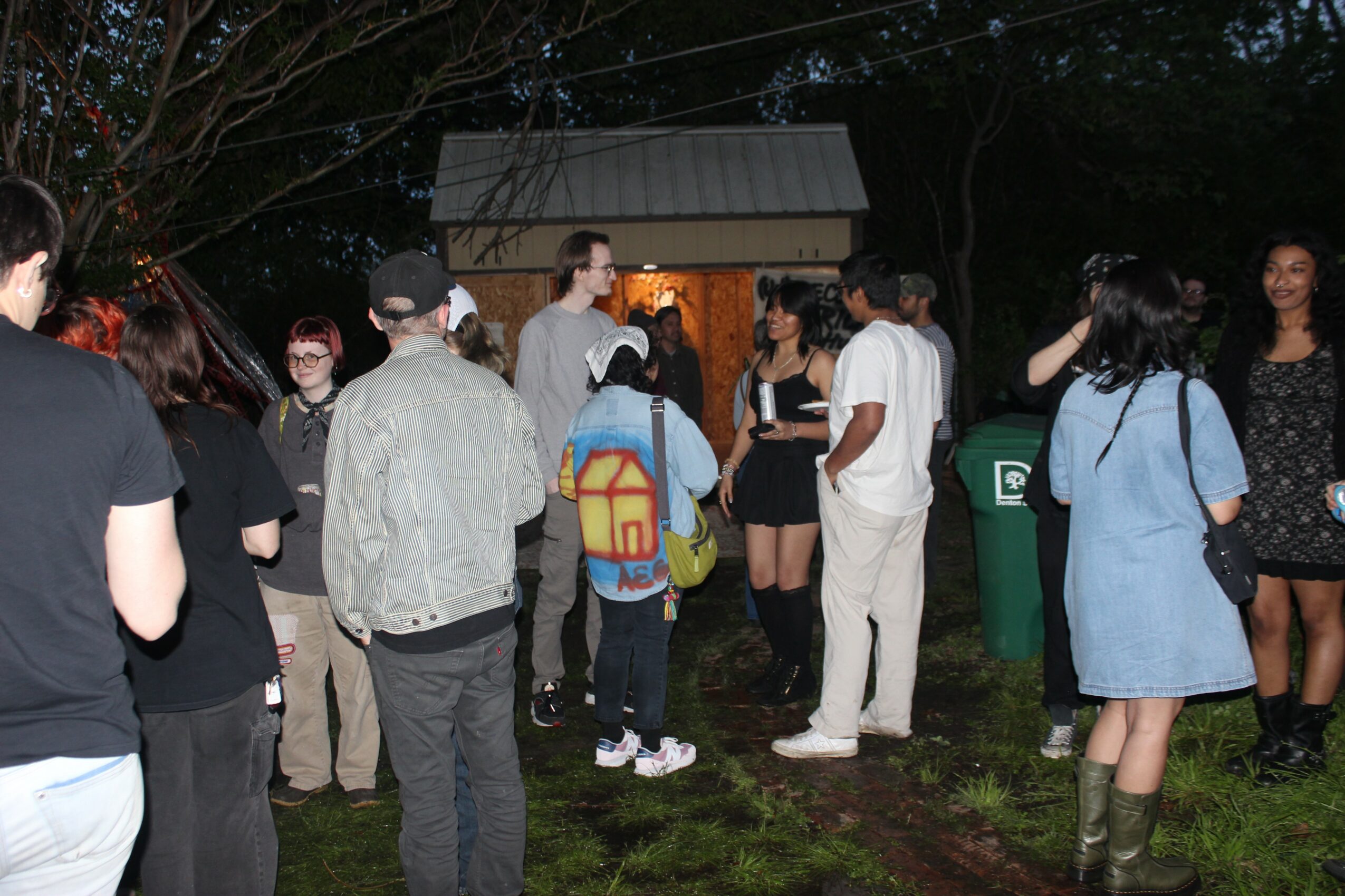Group of people gathered outdoors at night, some talking, some standing near a small building and a green trash bin.