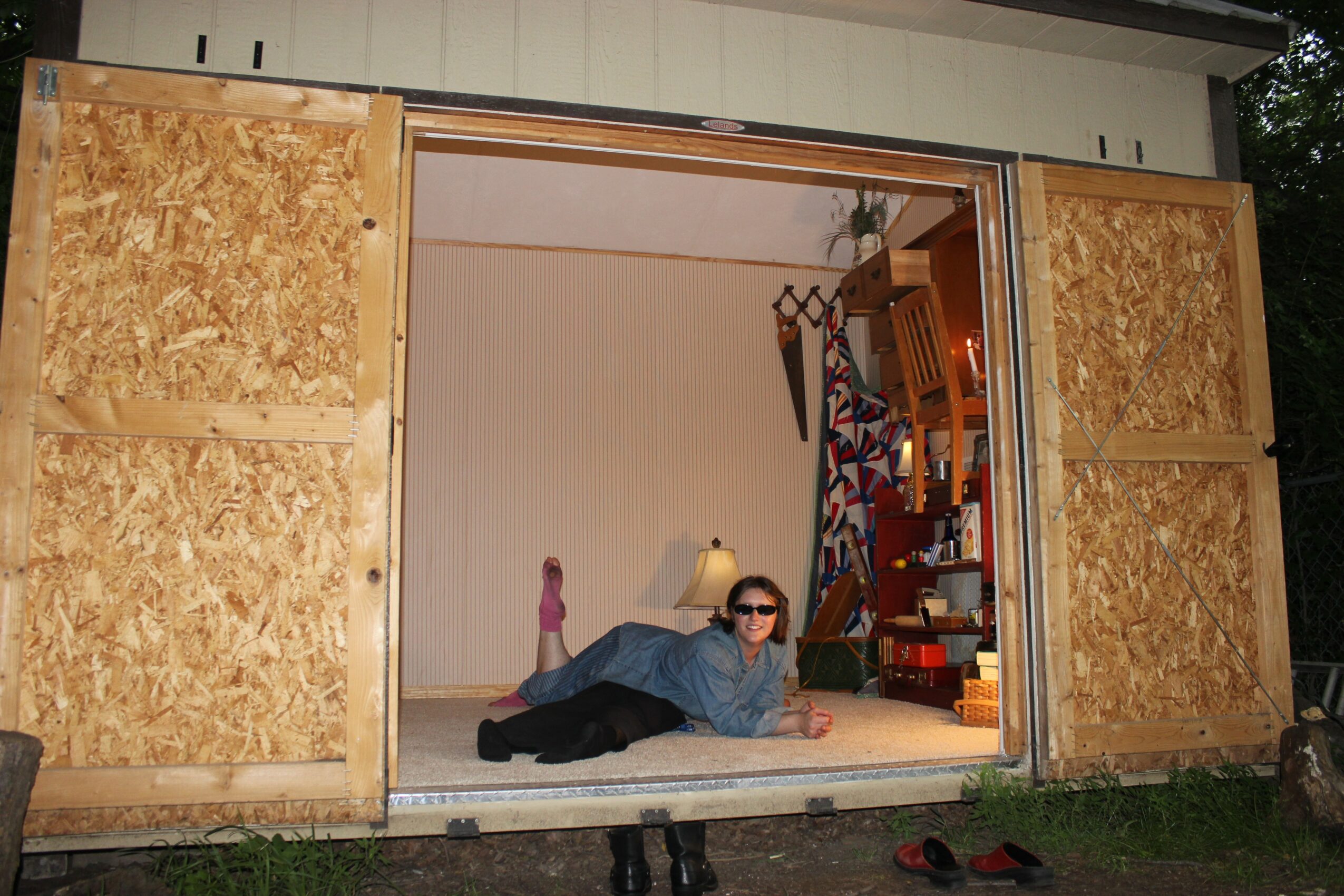 Person lying on the floor inside a shed with open doors, wearing sunglasses and a blue shirt, with a staircase inside.