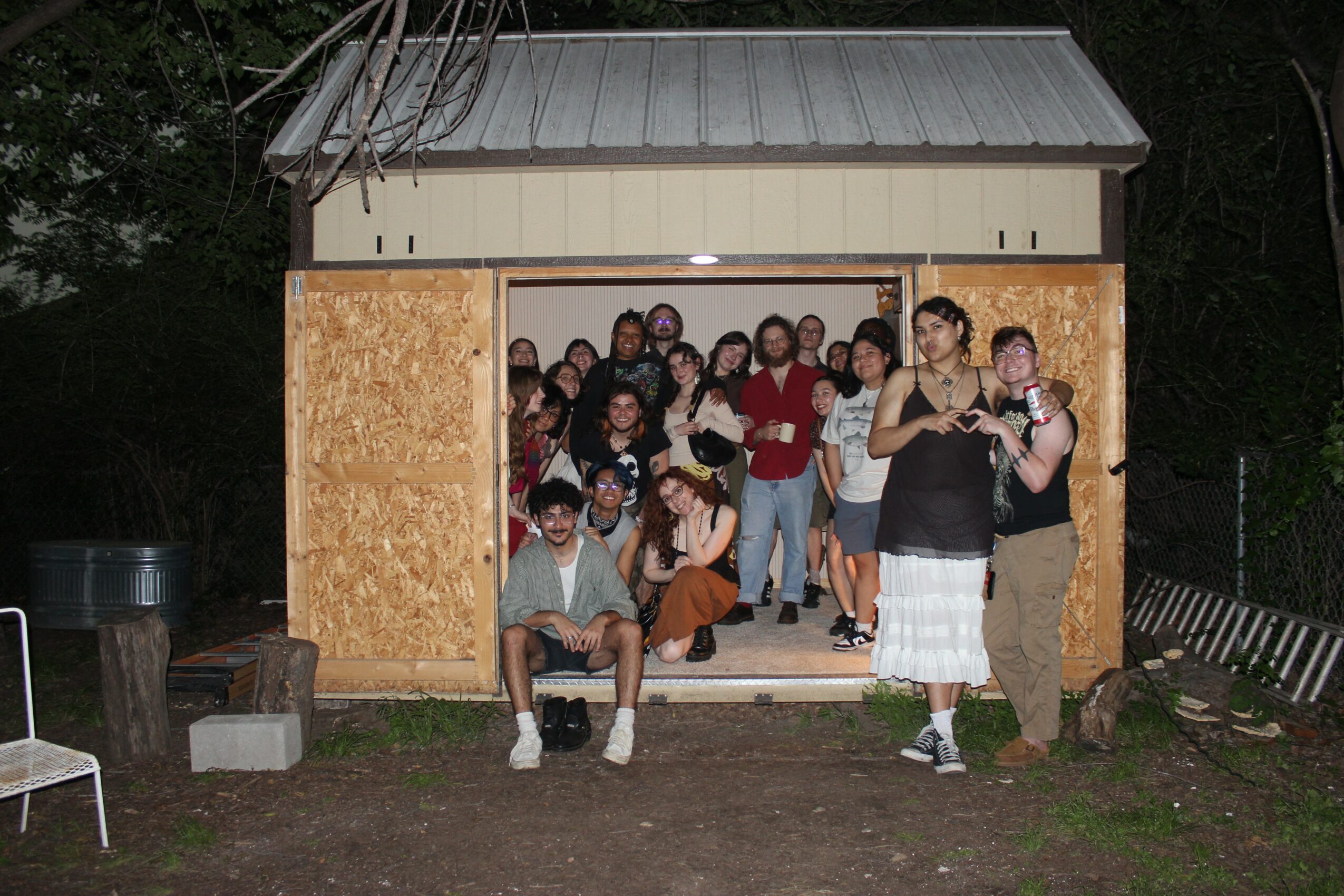 Group of people gathered inside a small wooden shed at night, some sitting and some standing, with a person holding a child outside.
