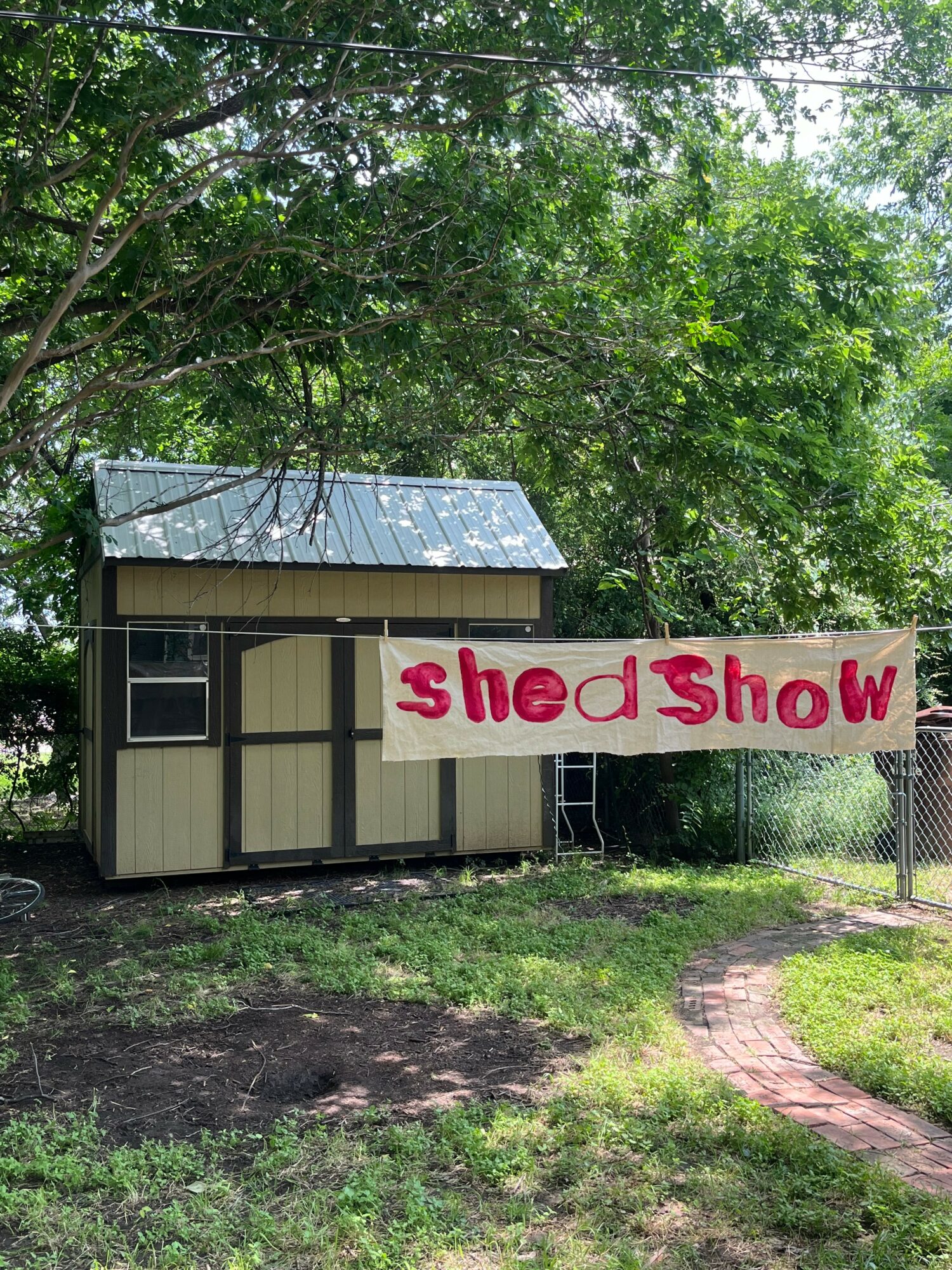 Small shed with a metal roof and a sign reading 'shed show' in pink letters, surrounded by trees and grass.