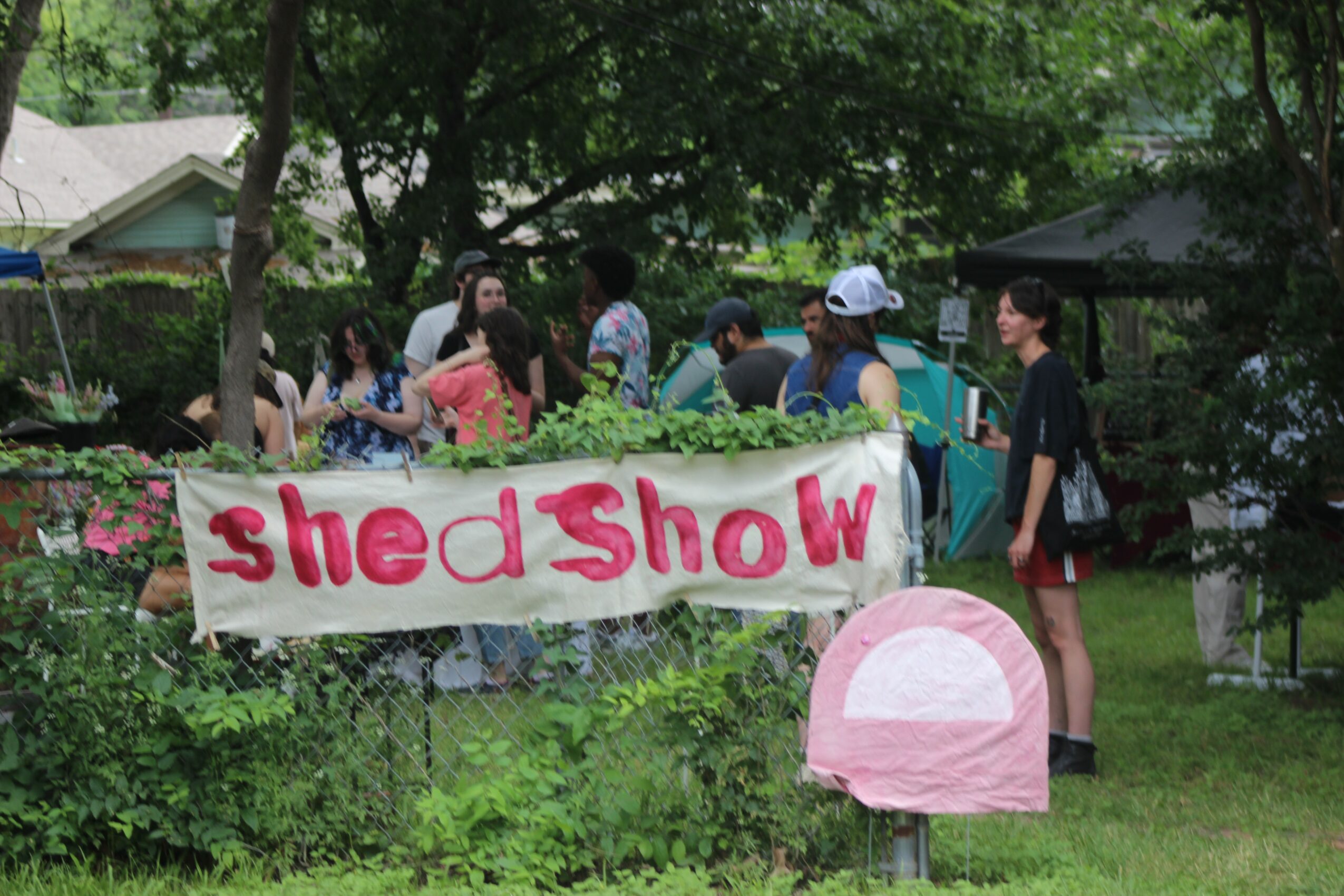 Group of children and adults outdoors behind a banner reading 'shed Show' in a garden with trees and greenery.