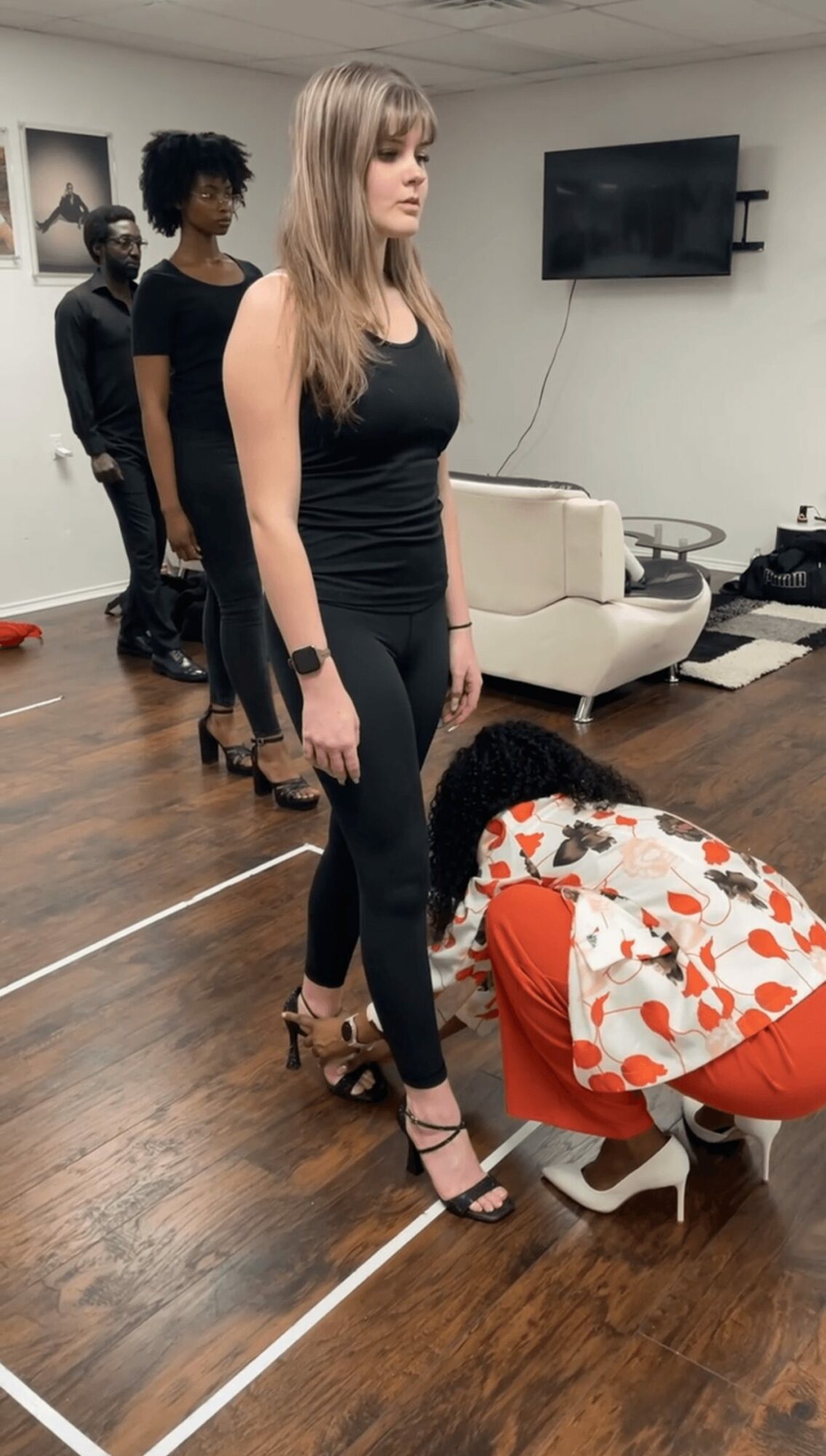 Four women in a room, one standing in front, others in line behind, with a person bending down in front of her.