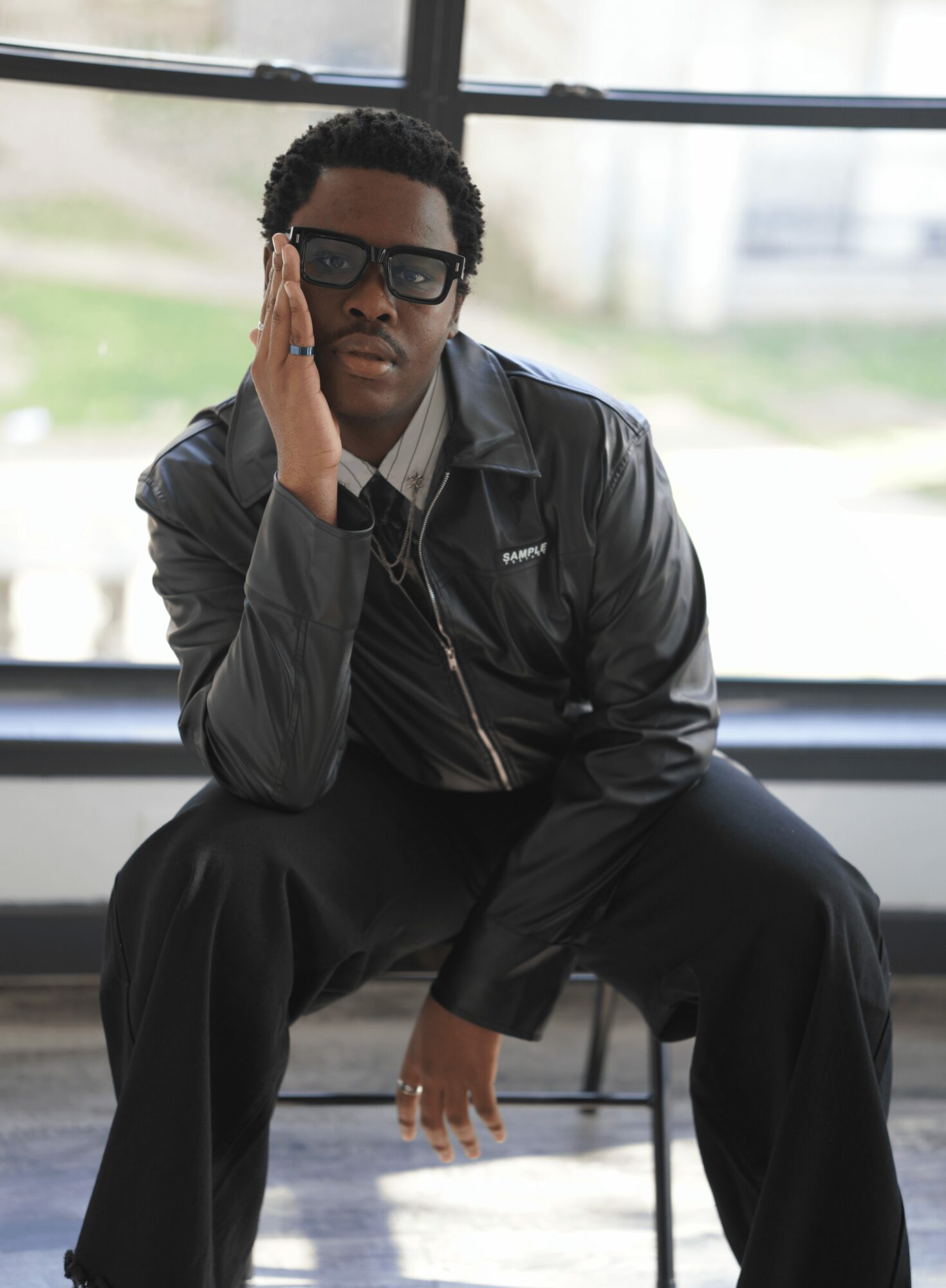 Person sitting indoors near large window, wearing sunglasses and a black jacket, resting their head on their hand.