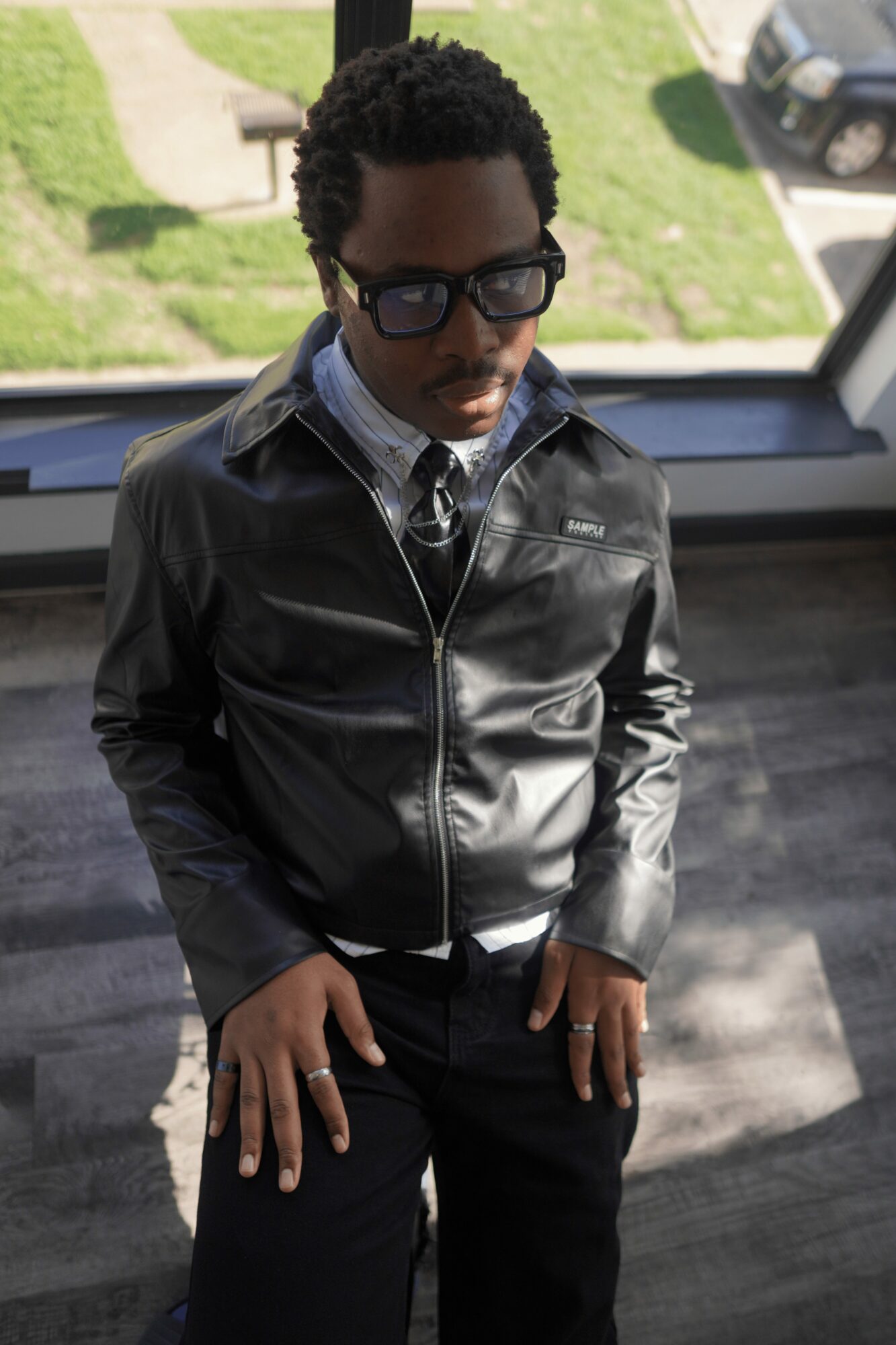 Young man with glasses and curly hair wearing a black leather jacket, standing indoors near a window with a view outside.