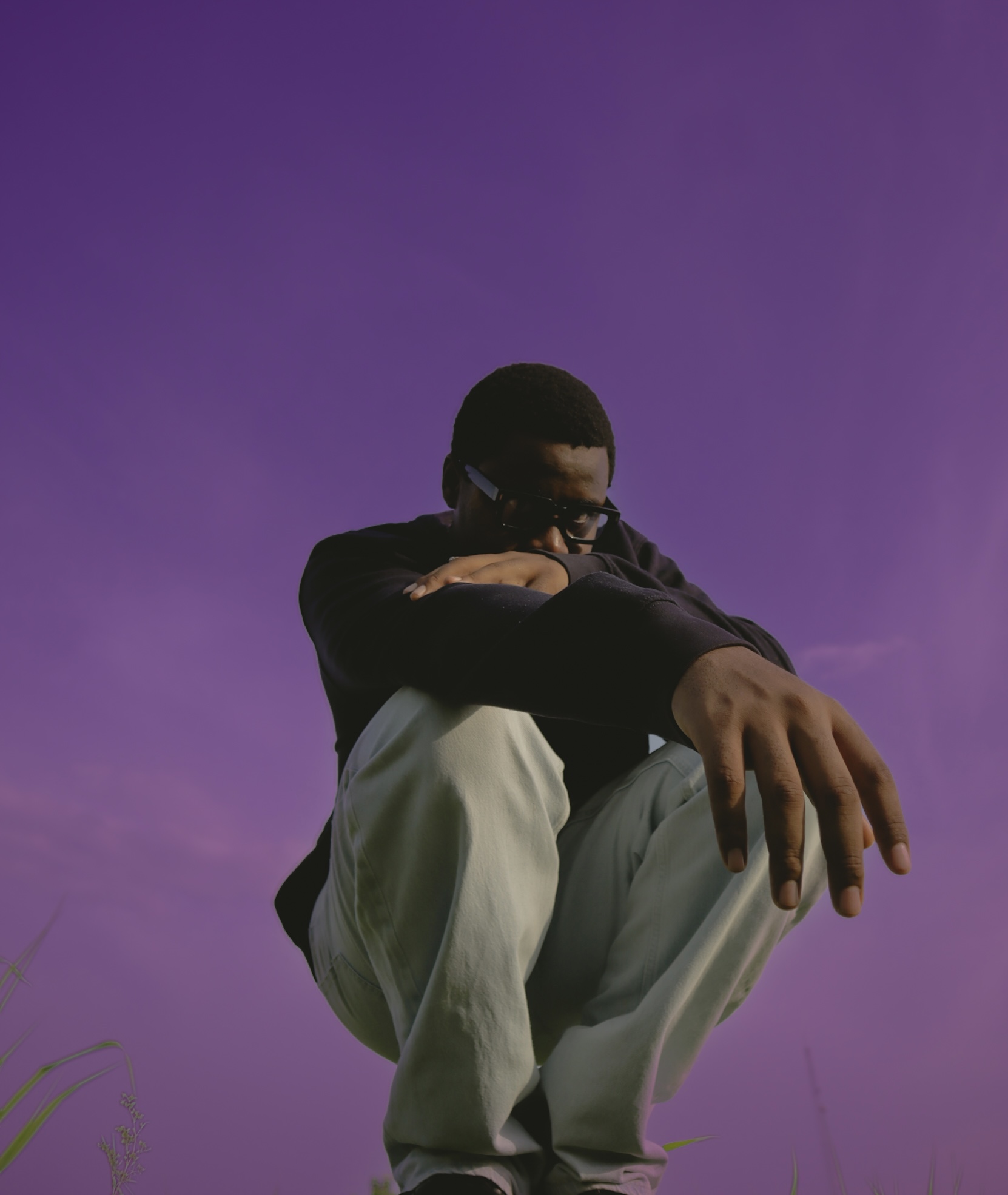 Person crouching with arms crossed, wearing glasses, against a purple sky background.