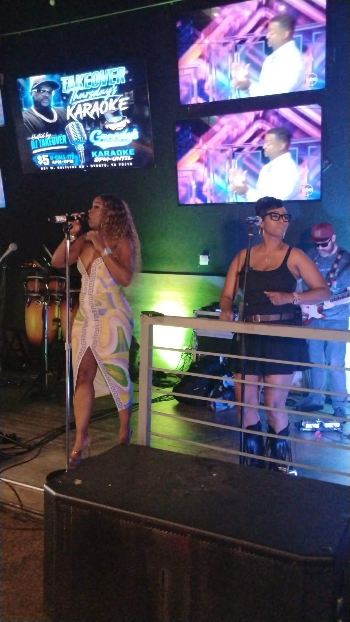 Two women perform on stage with screens displaying karaoke images behind them.