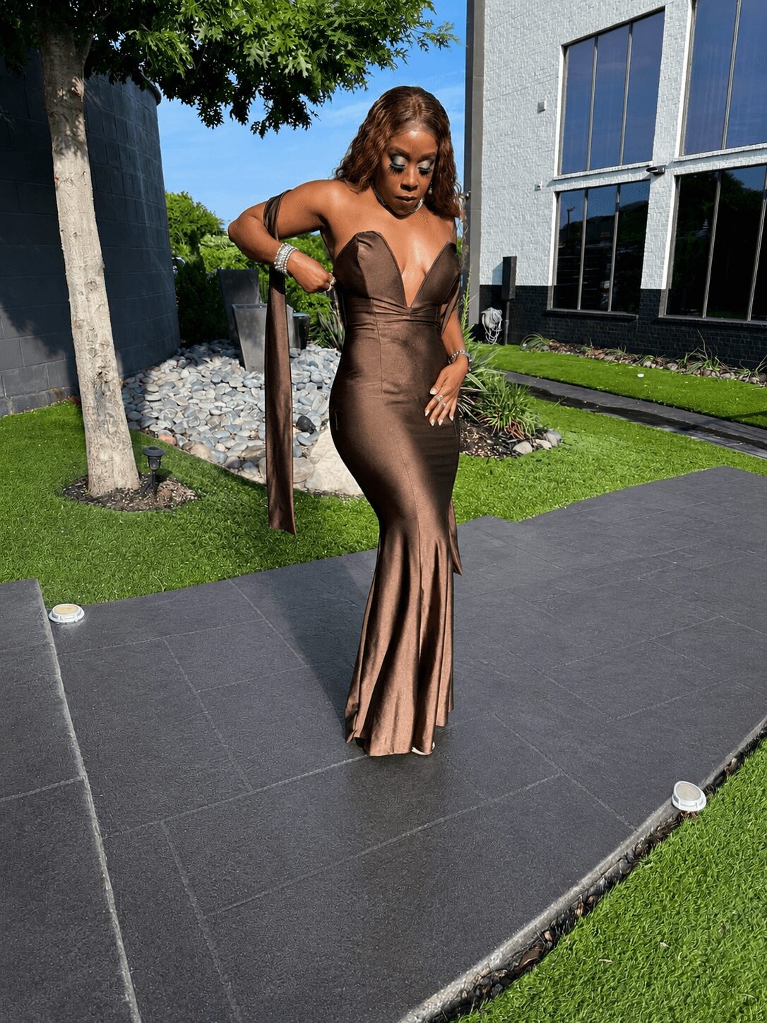 Woman in a brown gown standing outdoors on a paved area with grass and modern building in background.