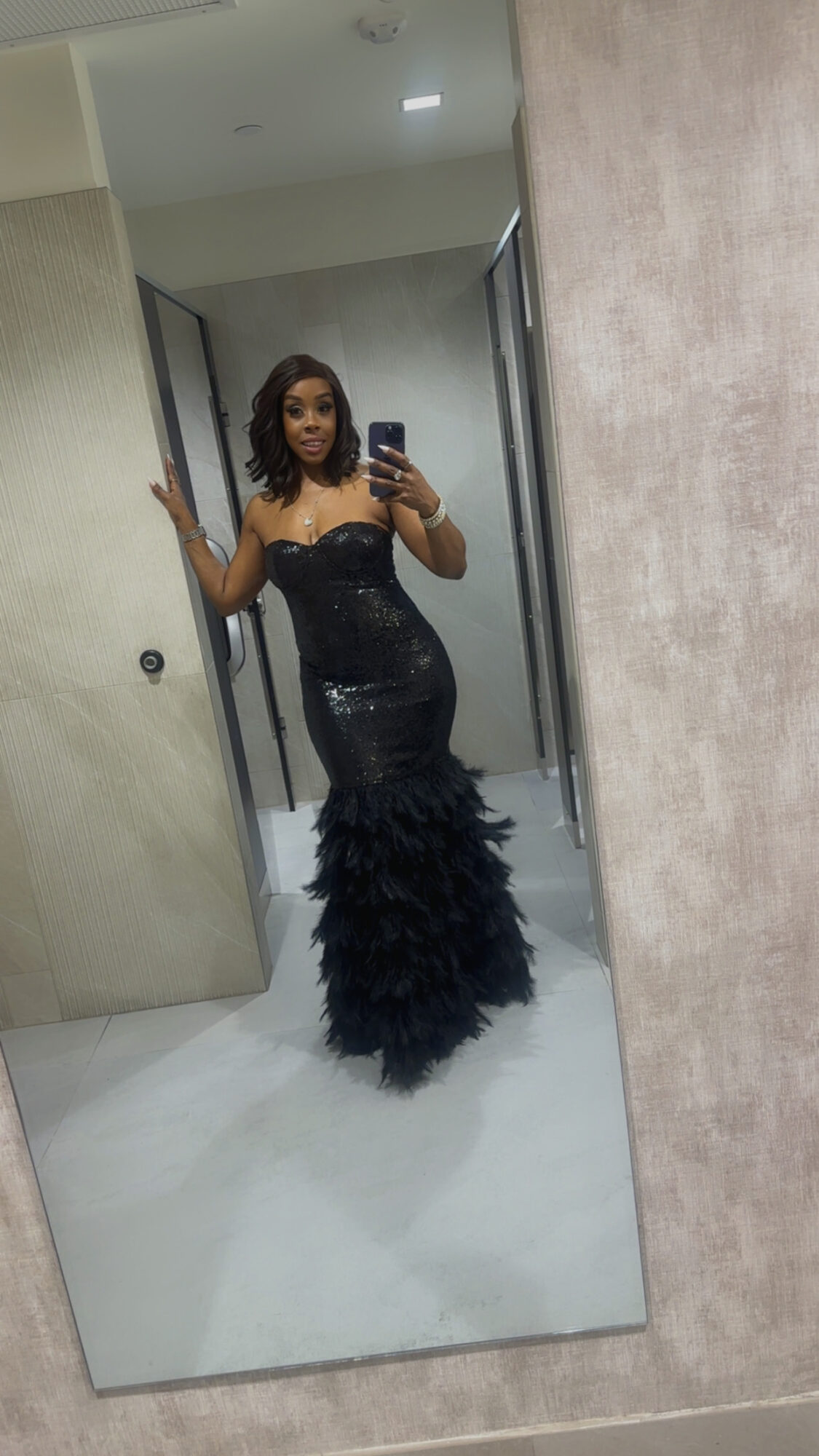 Woman in a black, strapless, feathered gown taking a mirror selfie in a fitting room.