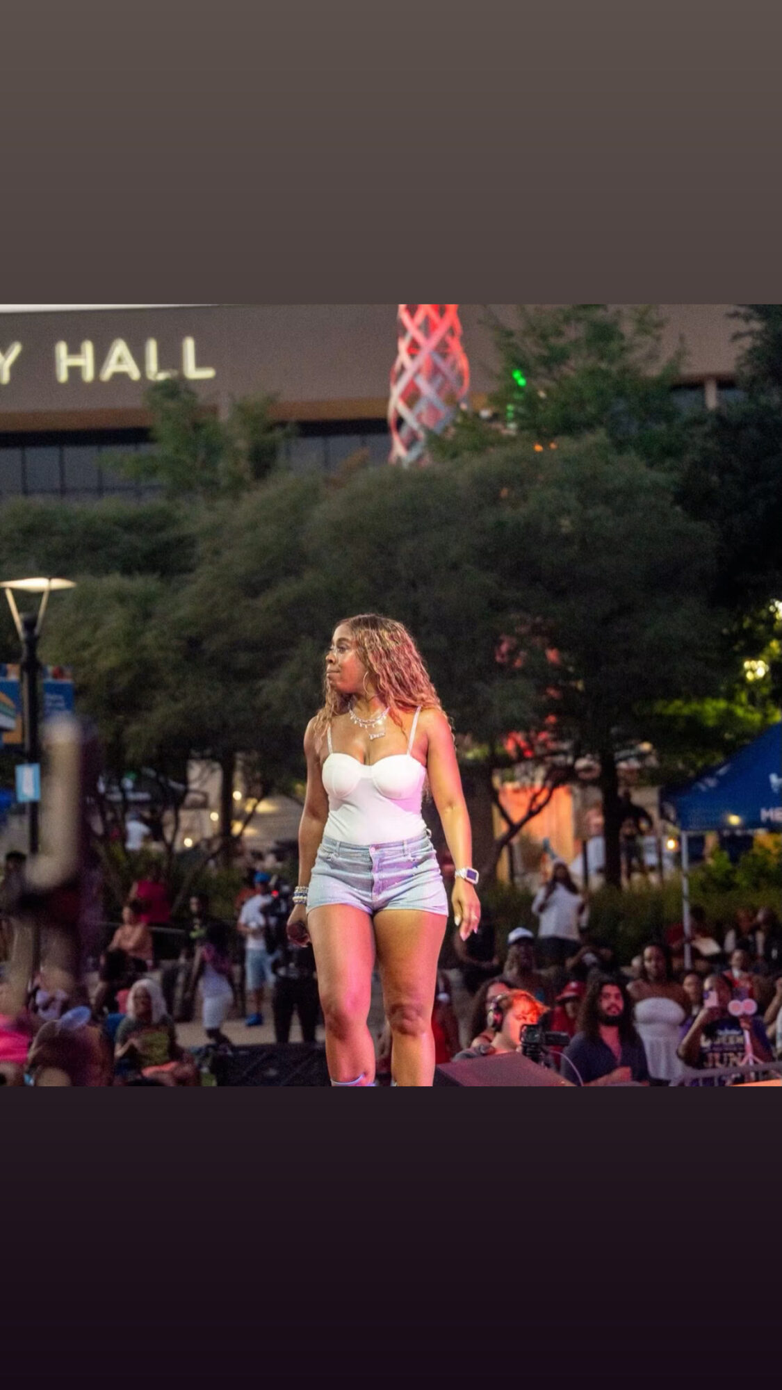 Young woman with curly hair in a white tank top and shorts on stage outdoors, audience in background, evening setting.