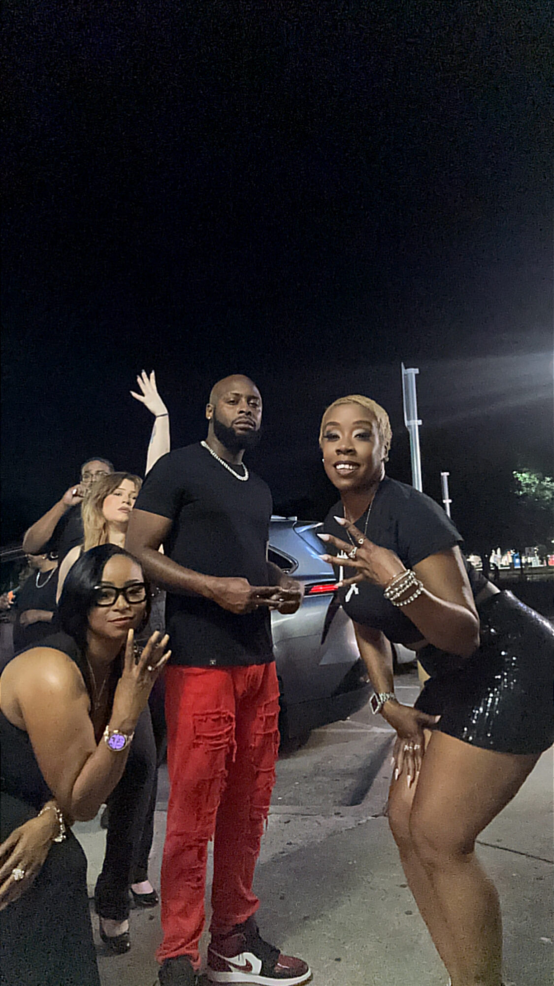 Group of five people posing outdoors at night, some making gestures, with a dark sky and parking lot in background.