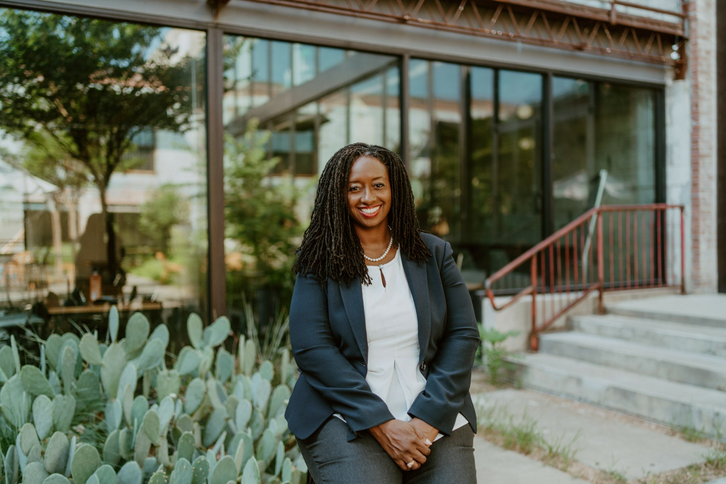 Smiling woman with curly hair in a navy blazer and white shirt standing outside near cactus plants, glass building background.