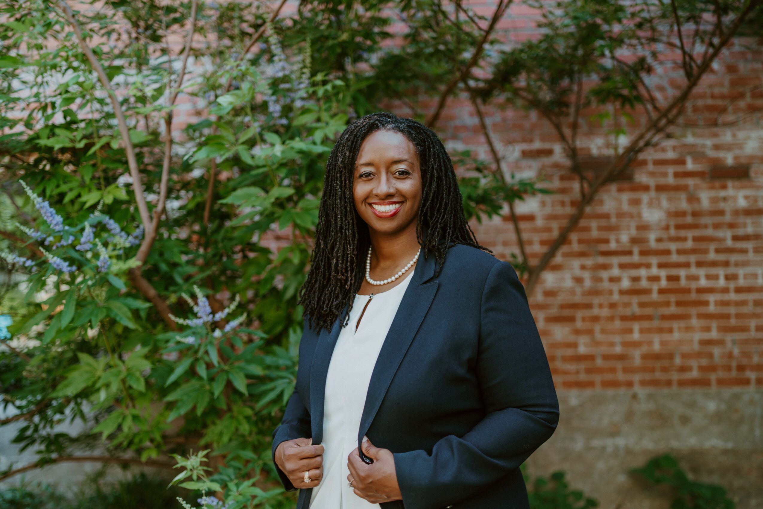 Smiling woman with long curly hair in a navy blazer and white top outdoors, brick wall and greenery behind her.
