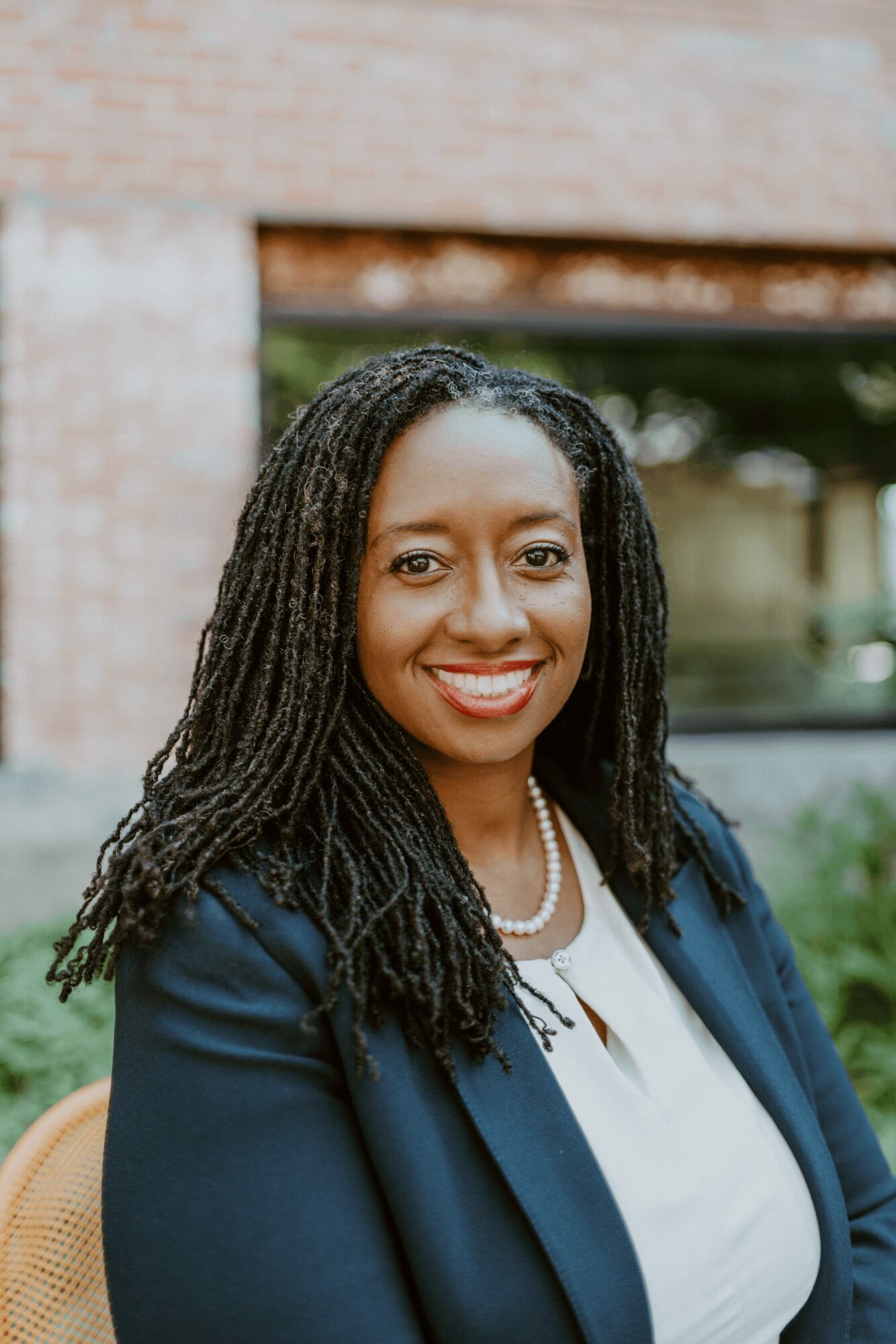 Smiling woman with long dreadlocks wearing a navy blazer and white top outdoors.