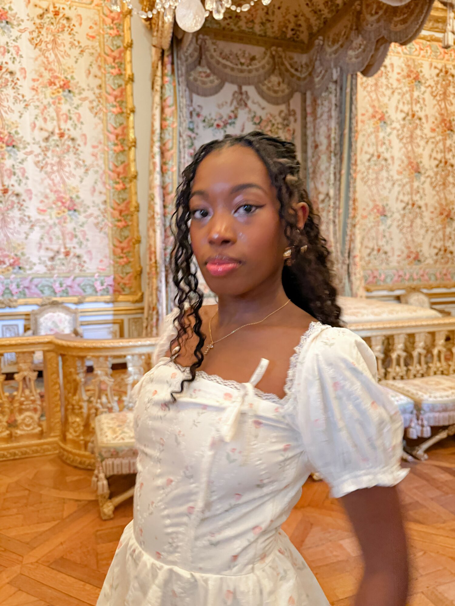 Young woman with curly hair in a white dress standing in a decorated room with ornate curtains and furniture.