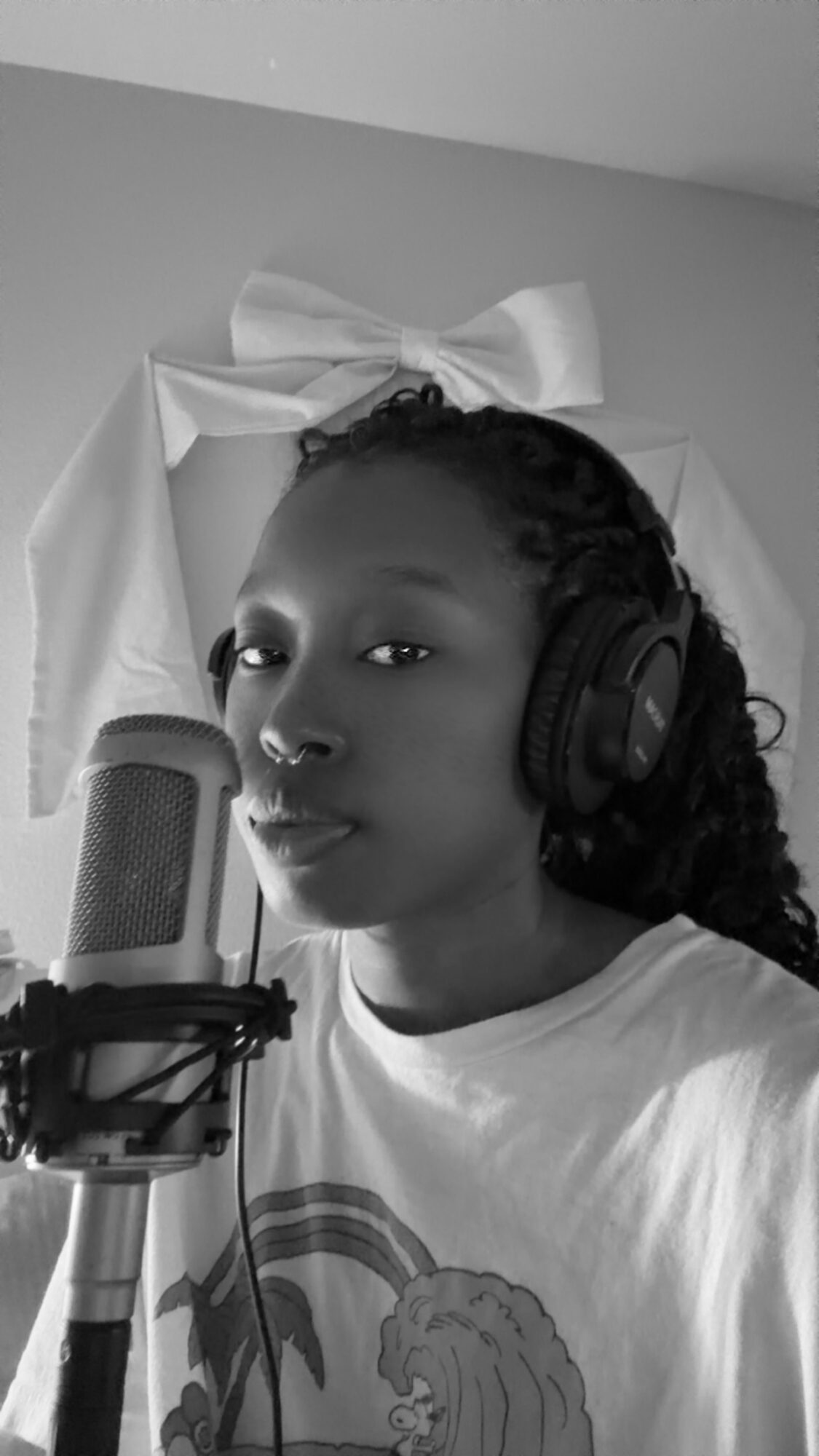 Young woman with curly hair wearing headphones, speaking into a microphone, in a room with a large bow decoration in the background.