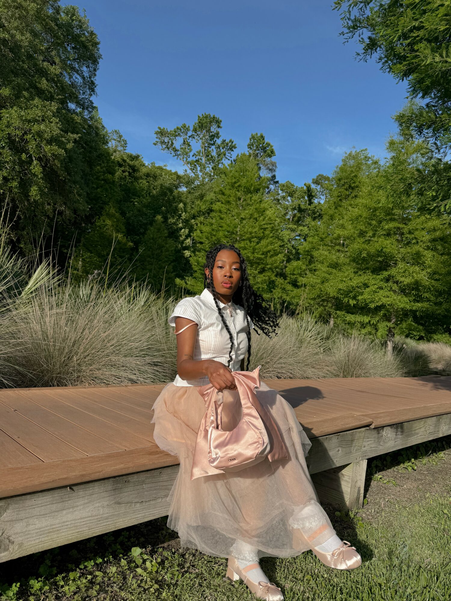 Young woman sitting on a wooden bench outdoors, surrounded by green trees and grass, with a blue sky overhead.