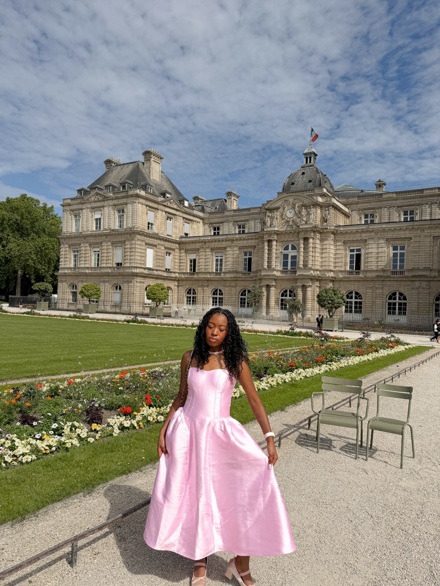 Young woman in pink dress standing in front of historic building with garden and benches, under partly cloudy sky.
