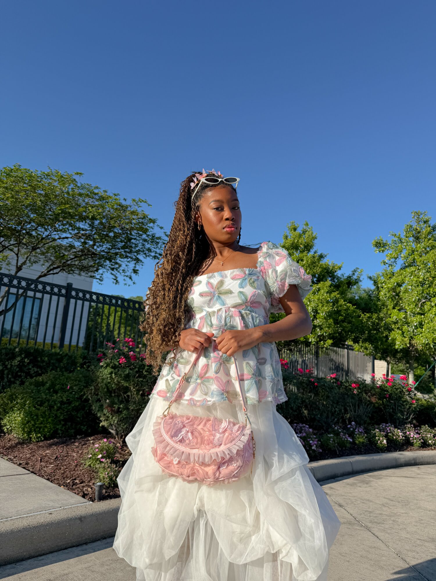 Young woman with long hair and sunglasses, wearing a floral top and white layered skirt, standing outdoors with trees and a fence behind her.