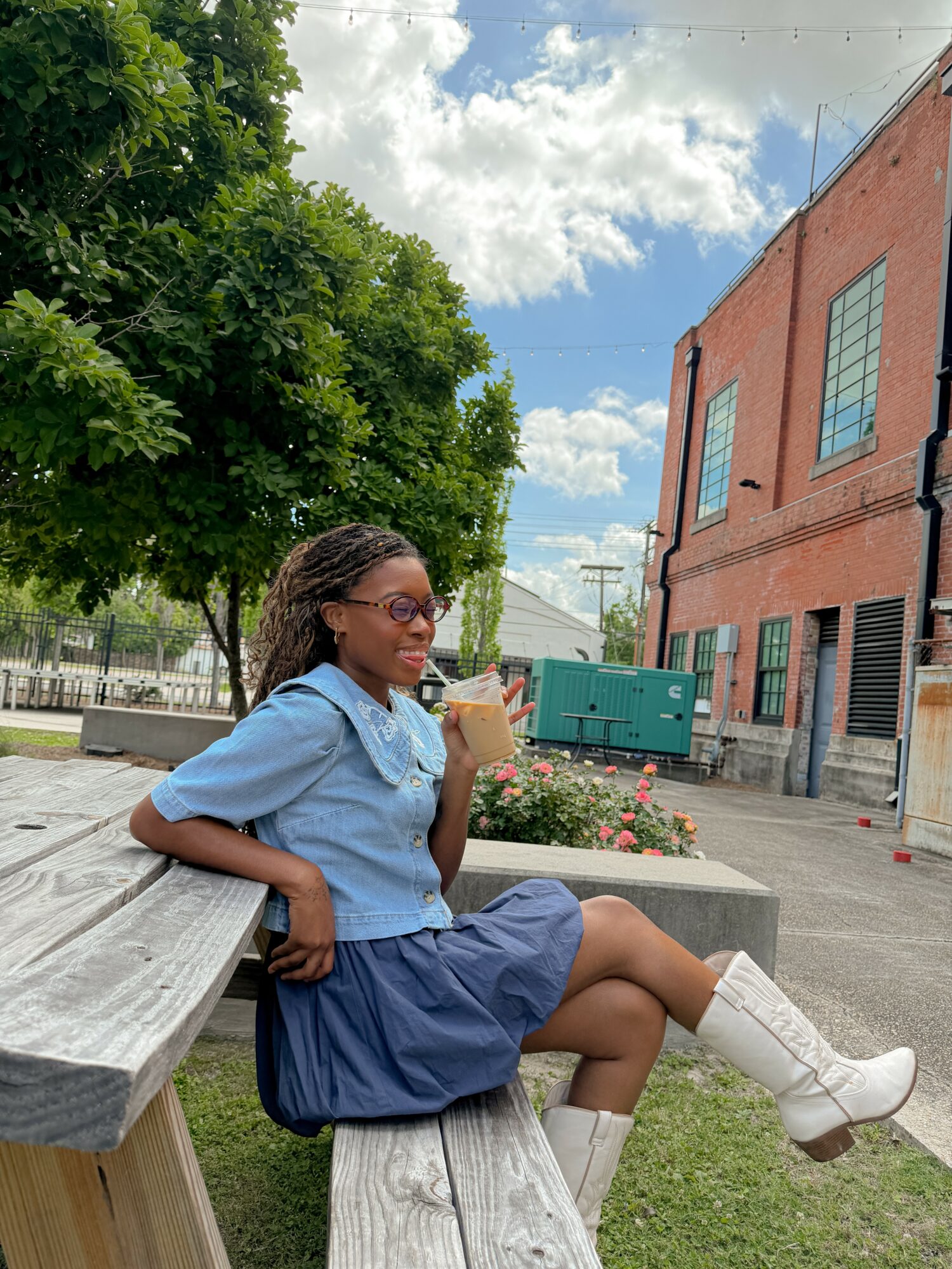 Young girl sitting on a bench outdoors, holding a drink, wearing glasses, a blue shirt, and a skirt, with trees and buildings in background.