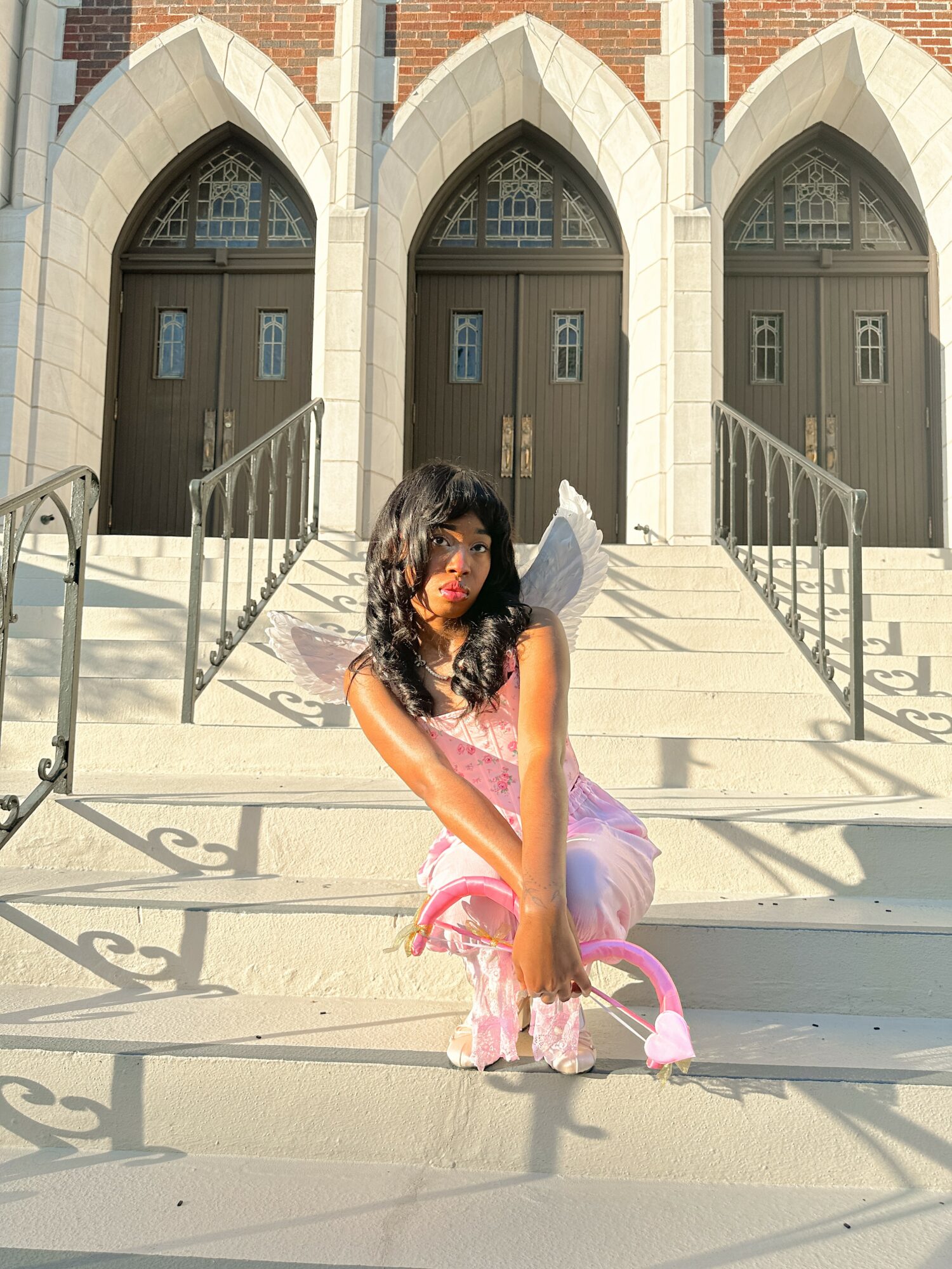 Child dressed as a fairy with wings and a pink tutu, sitting on stairs in front of a building with arched doors.
