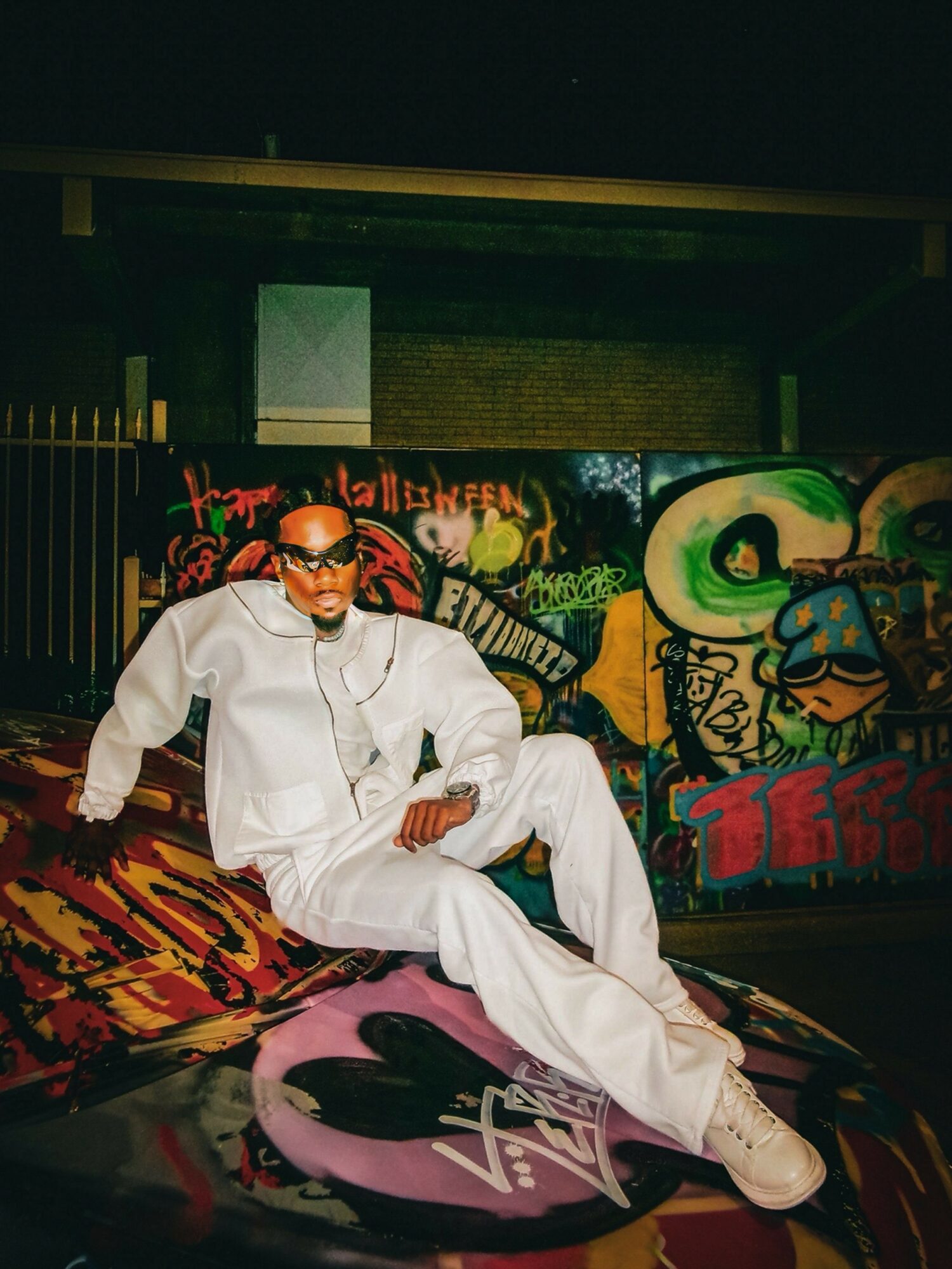 Man in white outfit and sunglasses sitting on graffiti-covered wall, with colorful street art behind him.