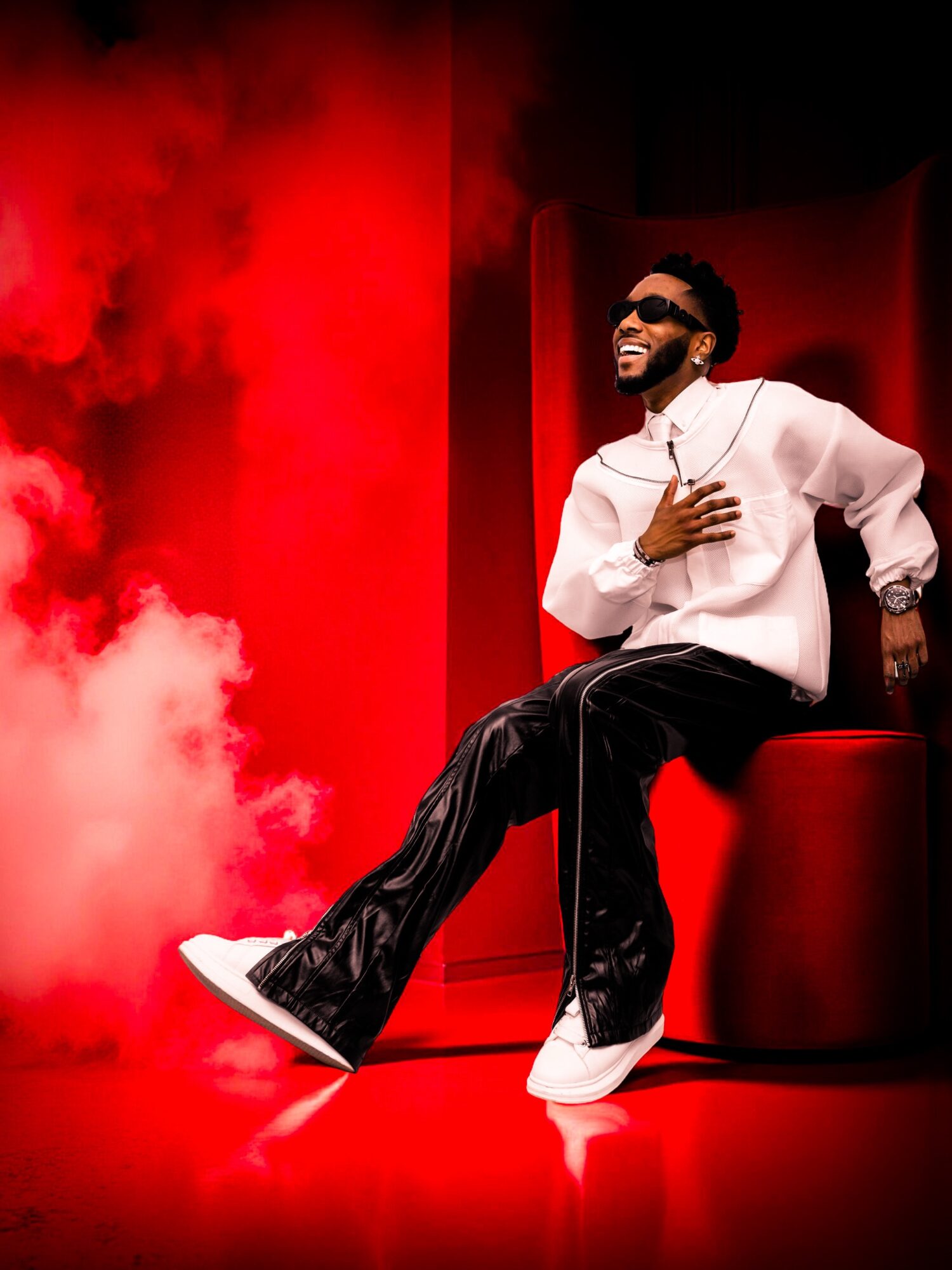 Person sitting on a red chair, smiling, wearing sunglasses, a white shirt, and black pants, with red background and smoke.