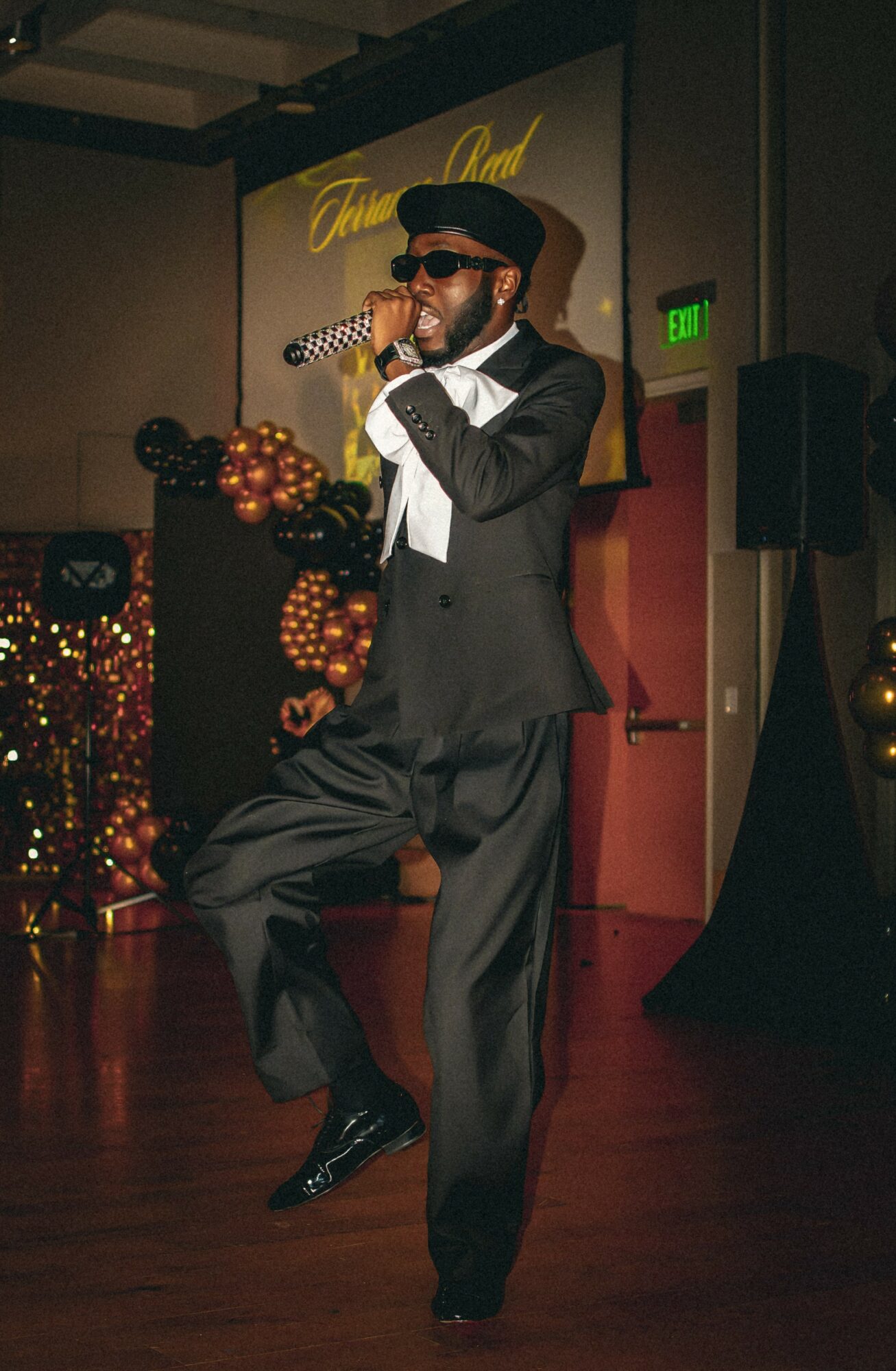 Man singing into a microphone on stage, wearing sunglasses, a hat, and a suit, with festive decorations in the background.