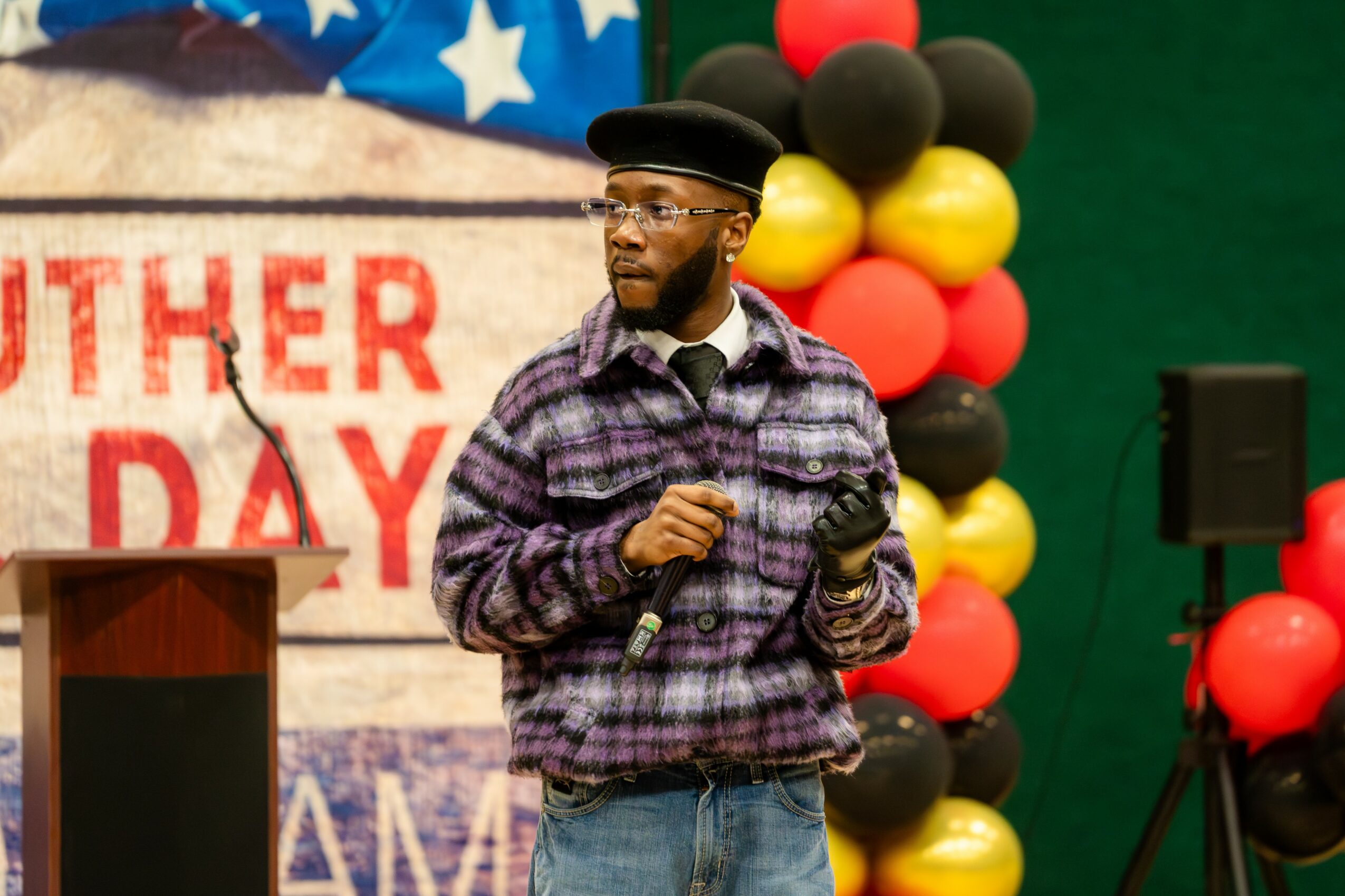 Man wearing a plaid shirt, glasses, and a black hat standing in front of a decorated background with balloons and a sign, holding a microphone.