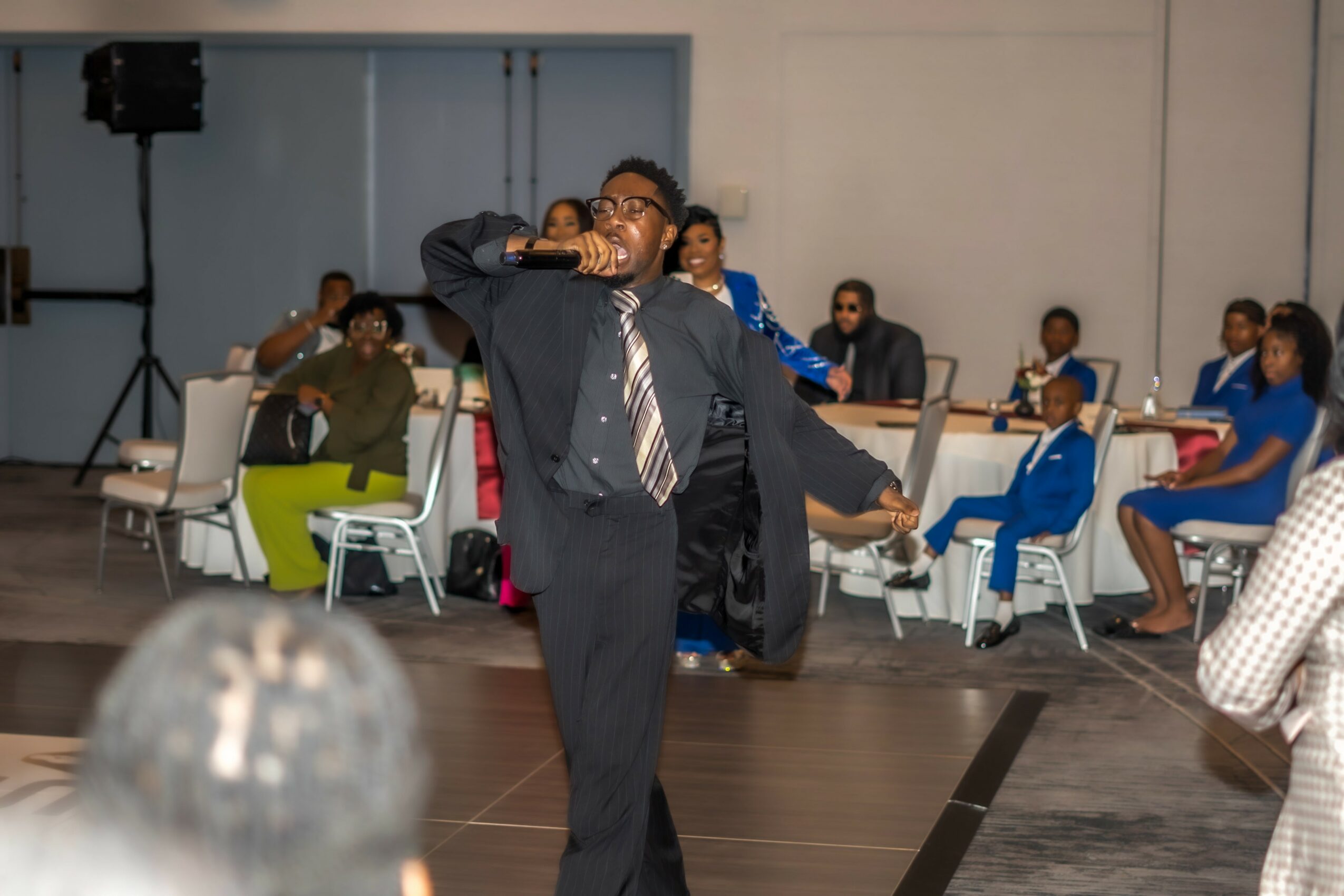 Person in suit drinking from a bottle while standing in a large room with seated people at tables.