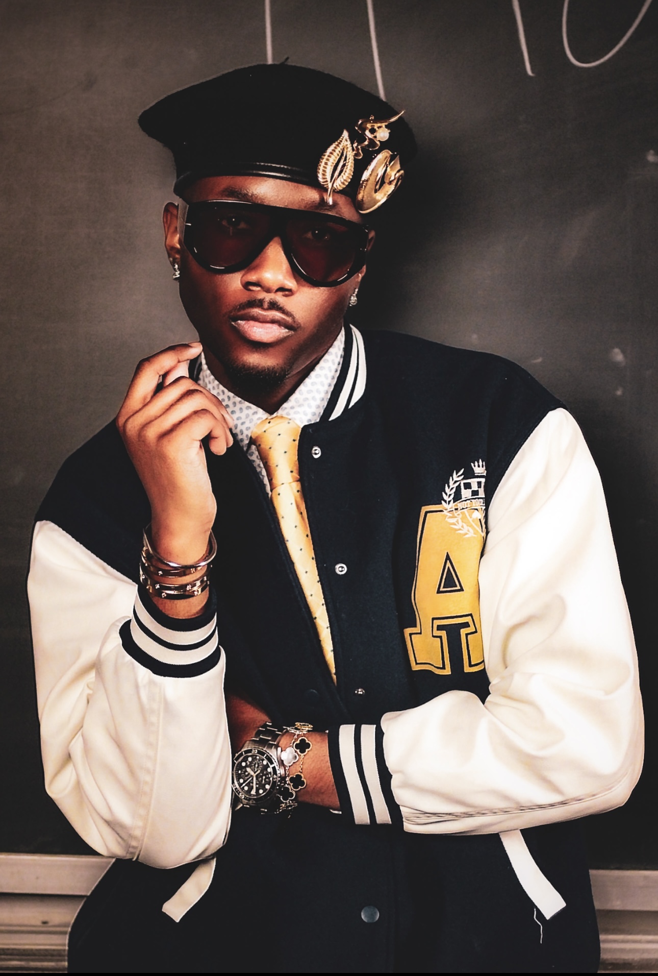 Young man wearing sunglasses, a cap, varsity jacket, and jewelry, posing with hand near face against dark background.