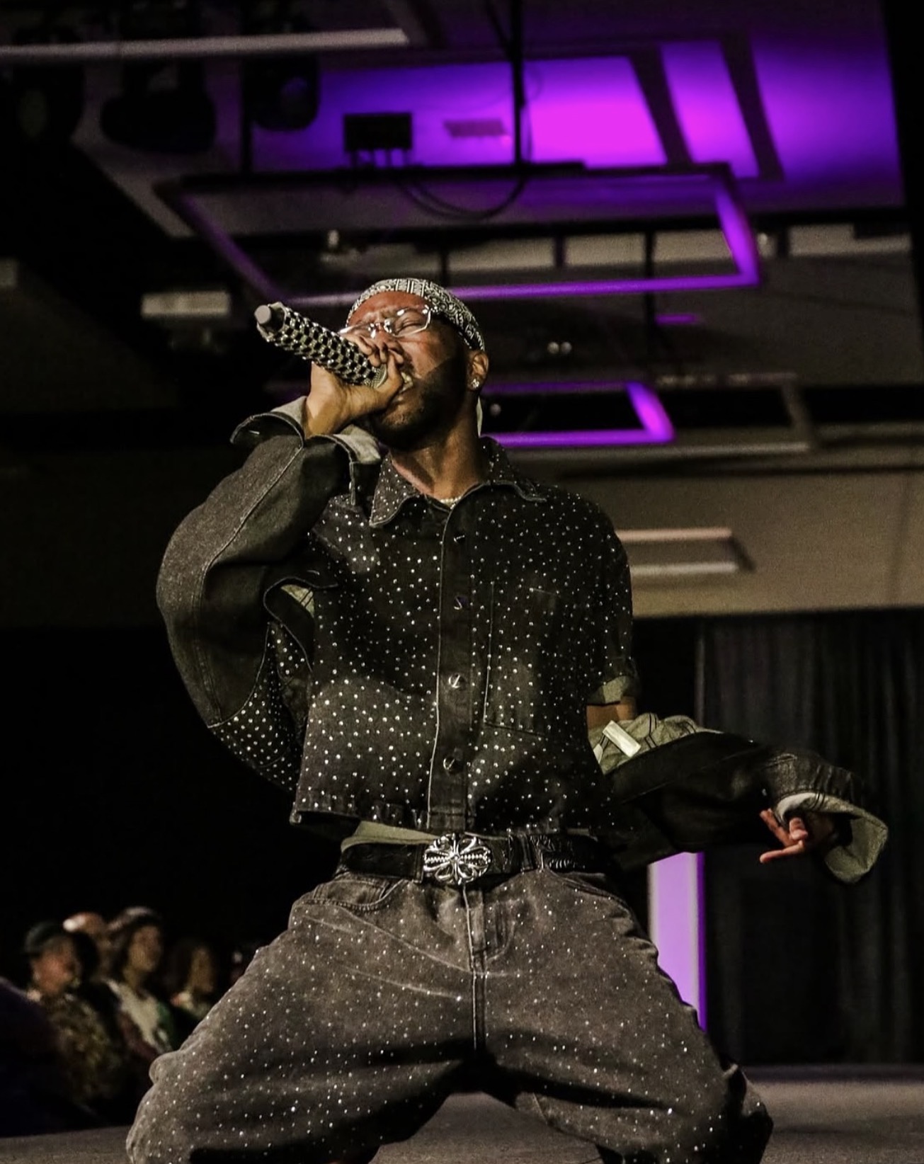 Performer singing into microphone on stage with purple lighting, wearing dark, sparkly clothing, and holding a jacket.