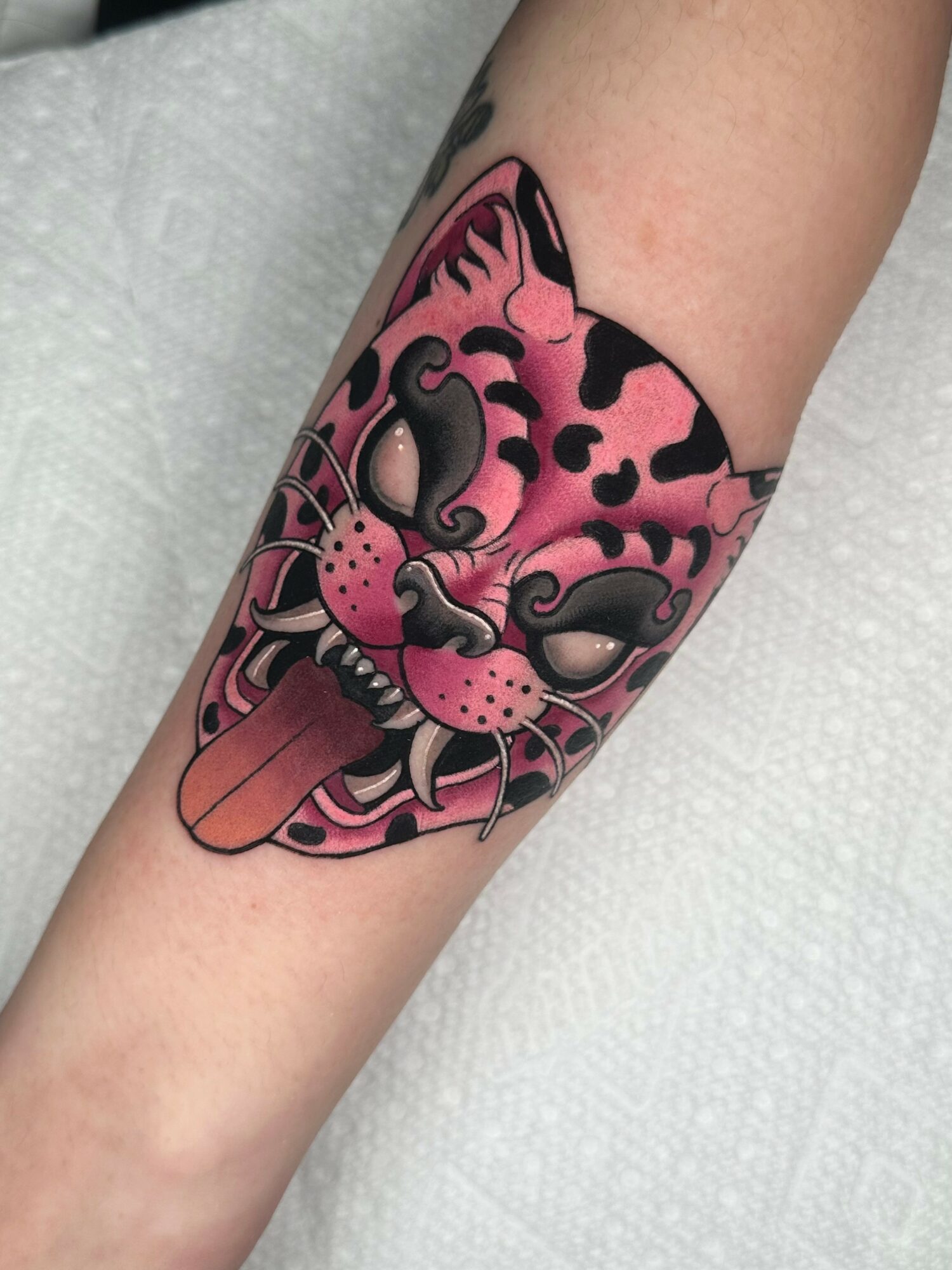 Tattoo of a pink leopard-print cat with a tongue sticking out, detailed face, and black accents.