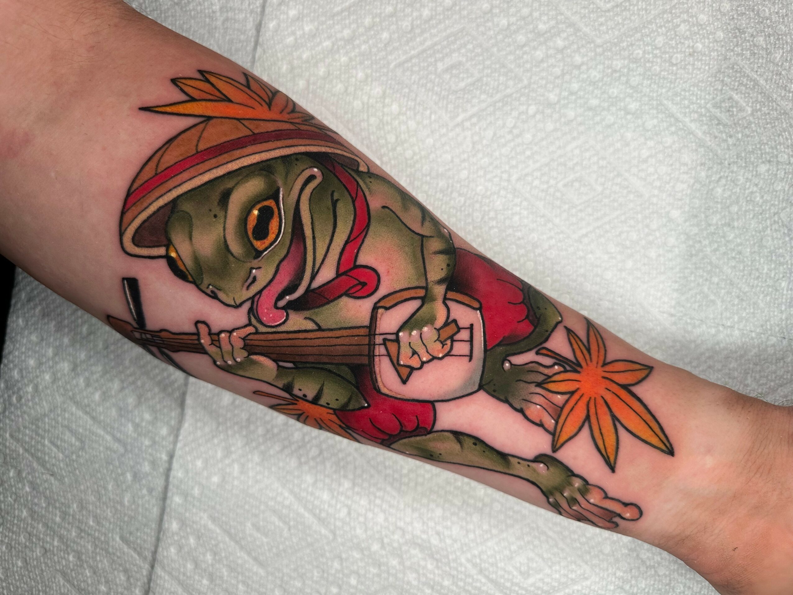 Colorful tattoo of a turtle wearing a hat, playing a guitar, with orange leaves, on a person's arm.